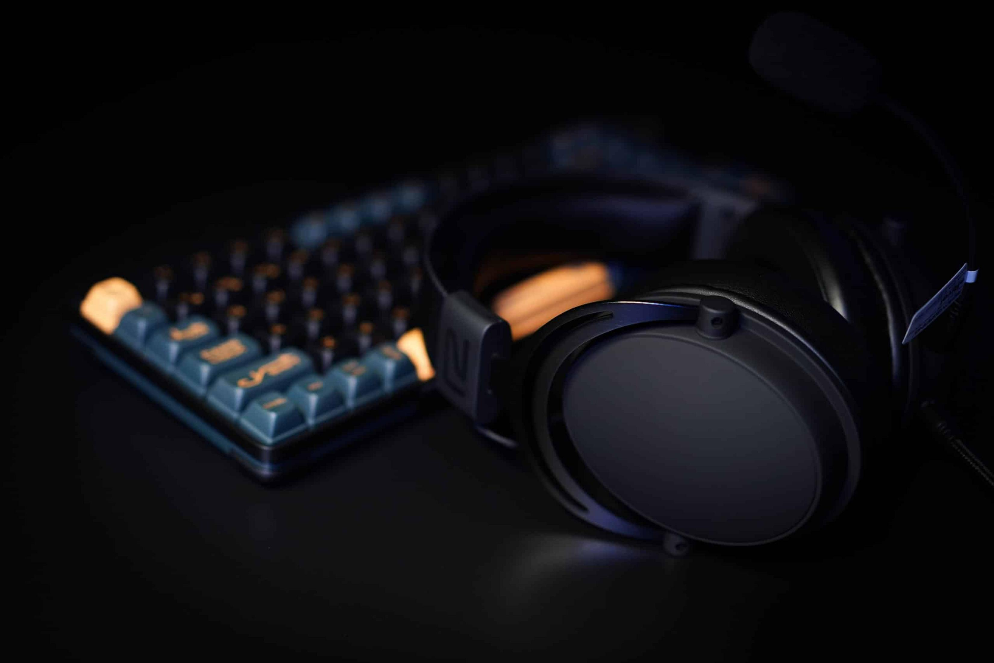 Schwarze Kopfhörer und eine blaue Tastatur mit orangefarbenen Tasten liegen auf dunklem Untergrund.