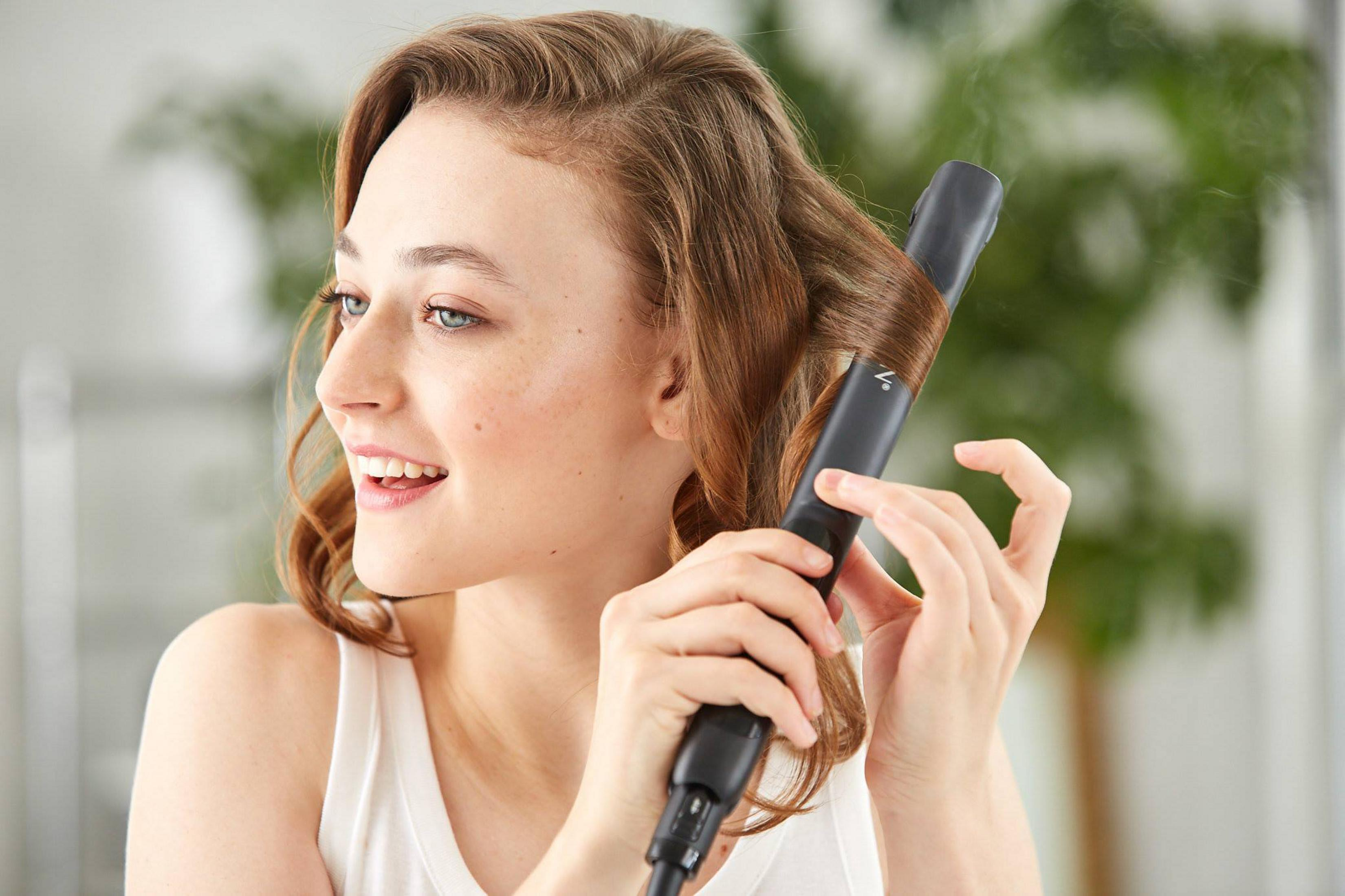 Kobieta z brązowymi włosami kręci je czarną lokówką. Unosi się para.