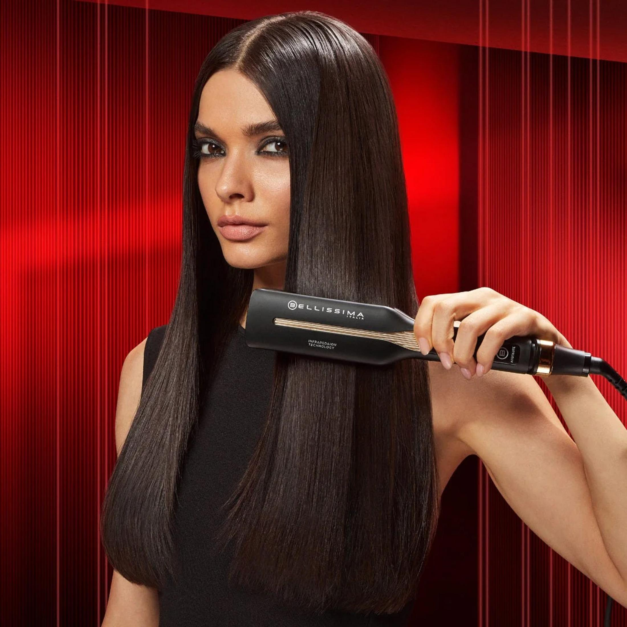 Kobieta z prostymi włosami trzyma czarną prostownicę na czerwonym tle.