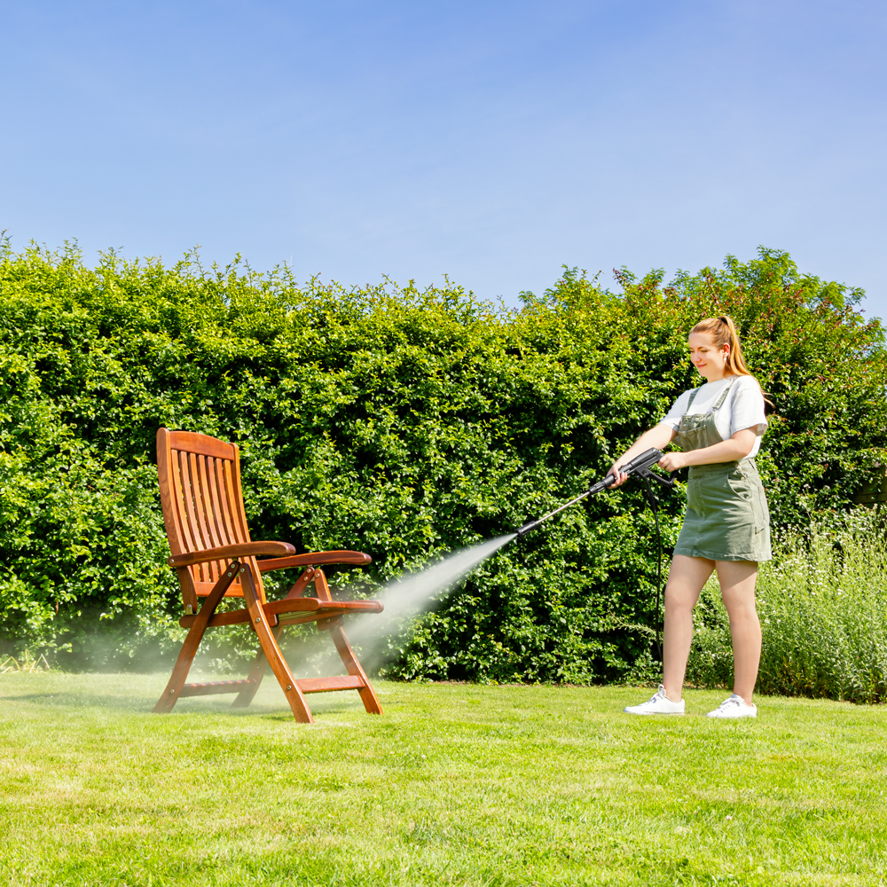Vrouw reinigt houten stoel met hogedrukreiniger in tuin.