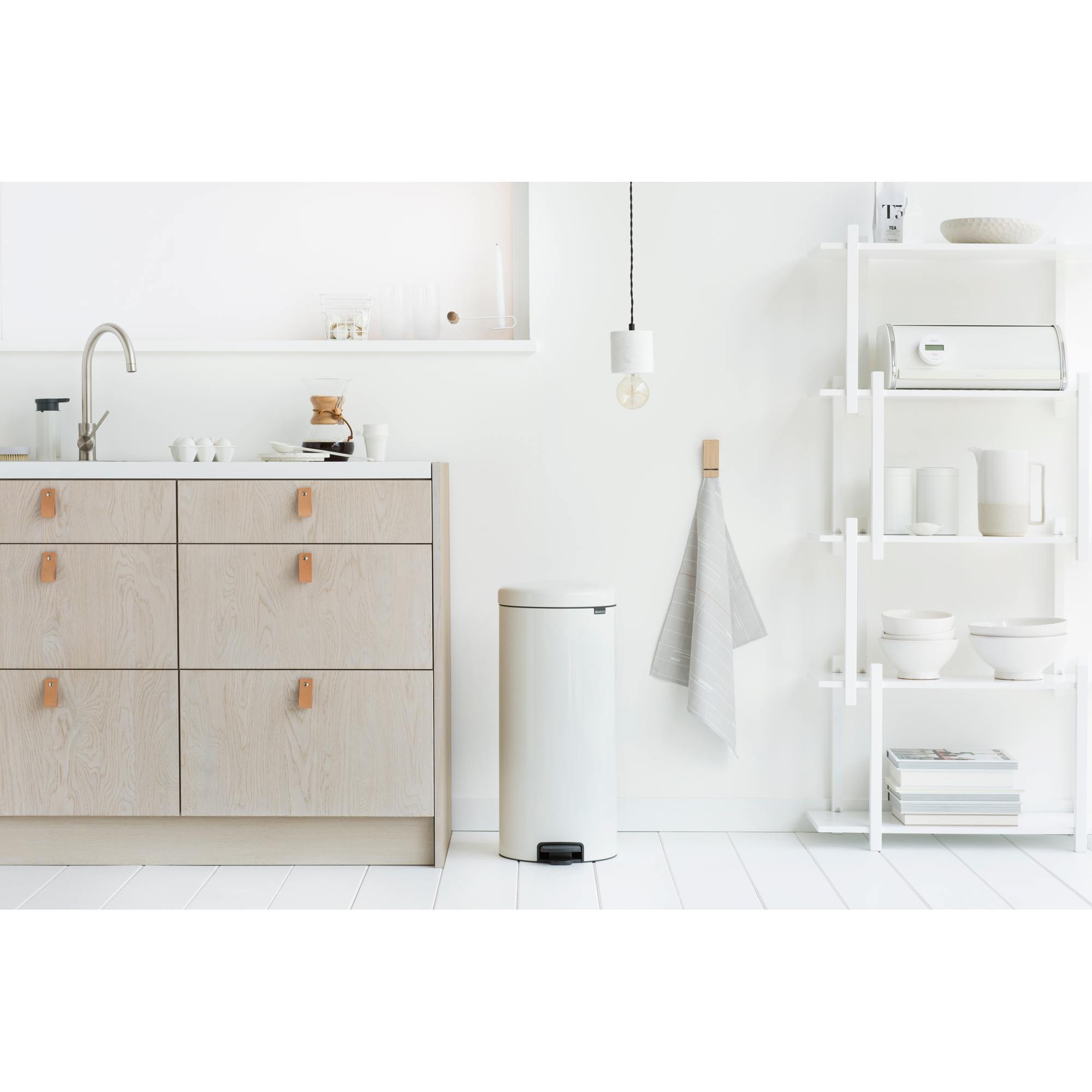 Een lichte, minimalistische keuken met lichte houten kasten, een witte prullenbak en planken.