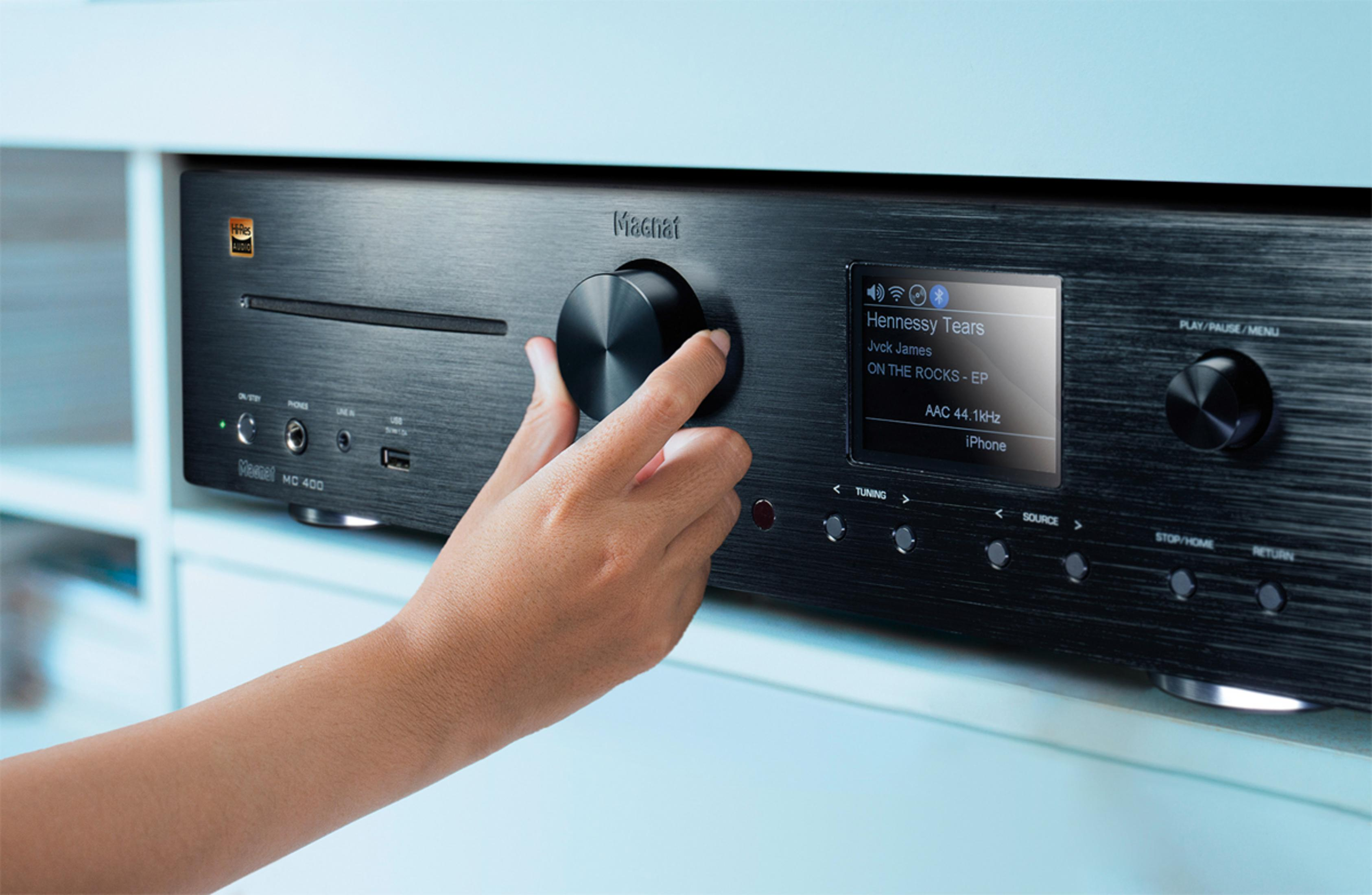 Eine Hand dreht an einem schwarzen Drehknopf eines silbernen Audiogeräts mit Display.