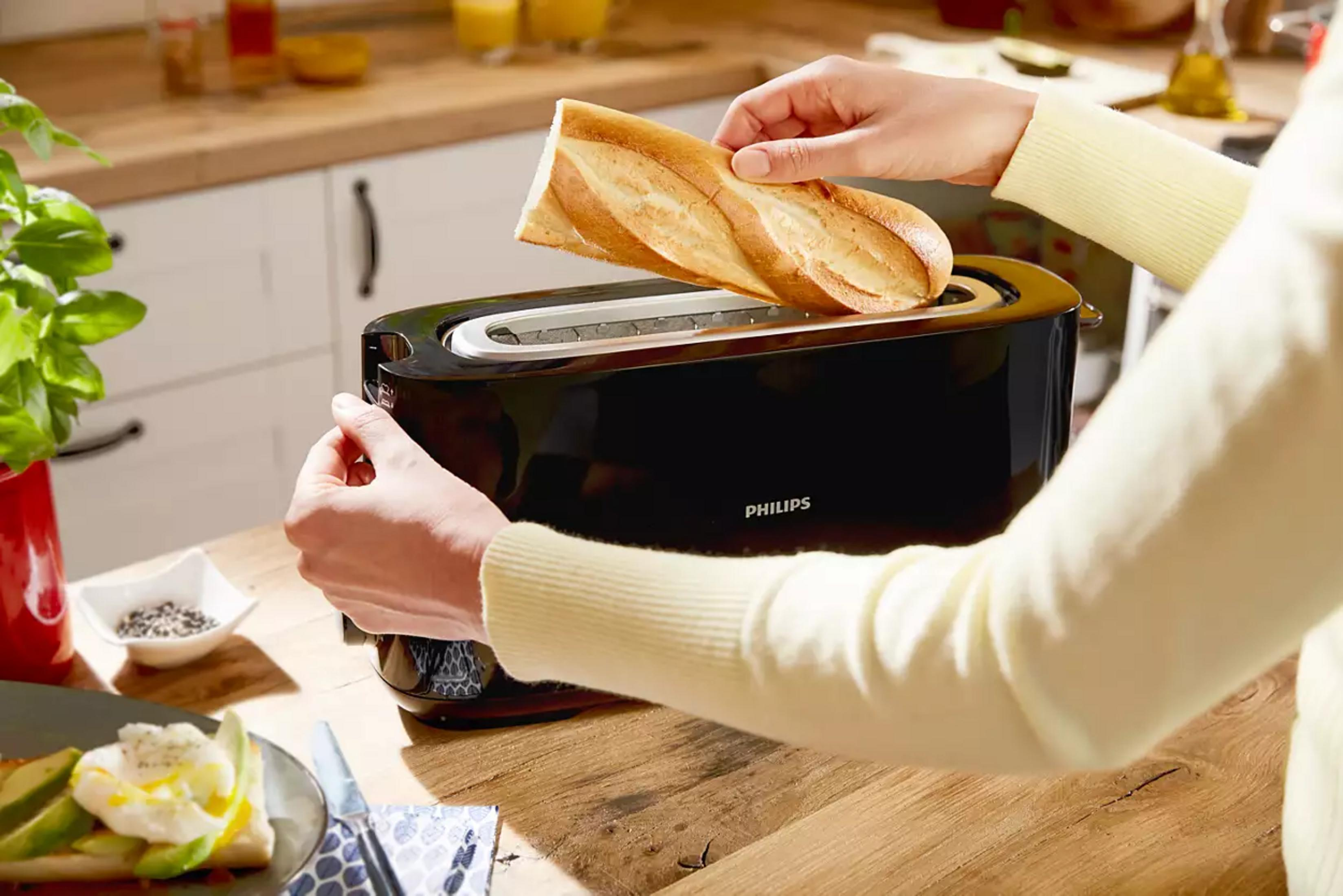 Una mano mette una baguette in un tostapane nero. A sinistra, una pianta di basilico.