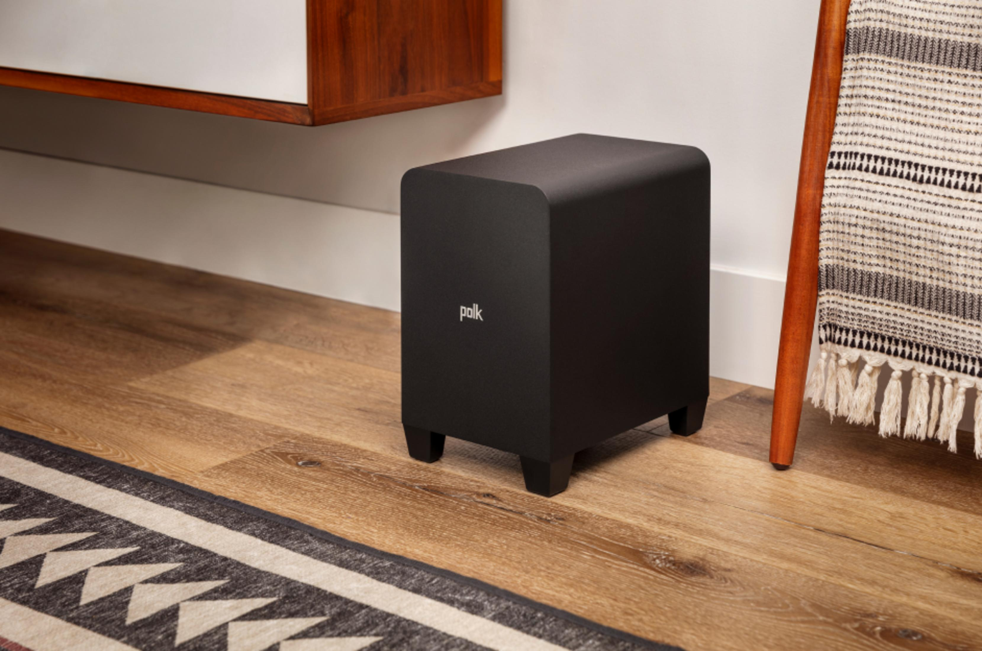 Czarny subwoofer Polk na drewnianej podłodze, obok dywanu i drewnianej nogi.