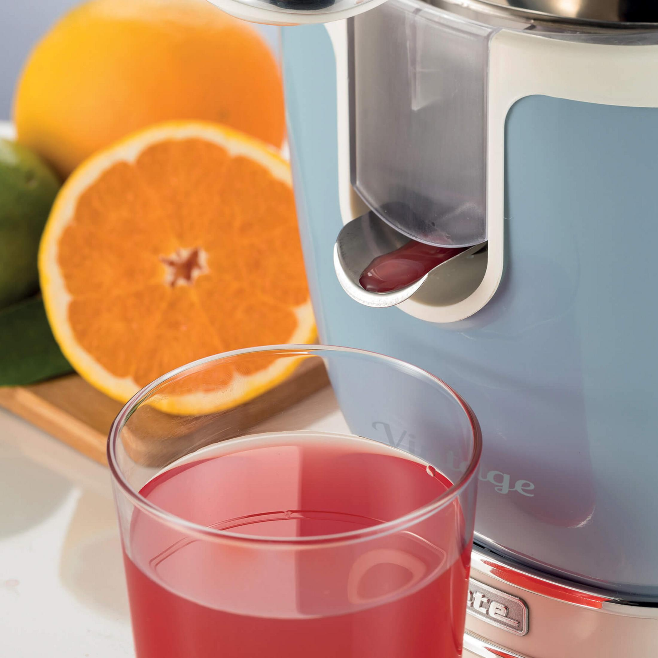 Ein Glas roter Saft steht neben einem Entsafter mit Orangen und Limetten.