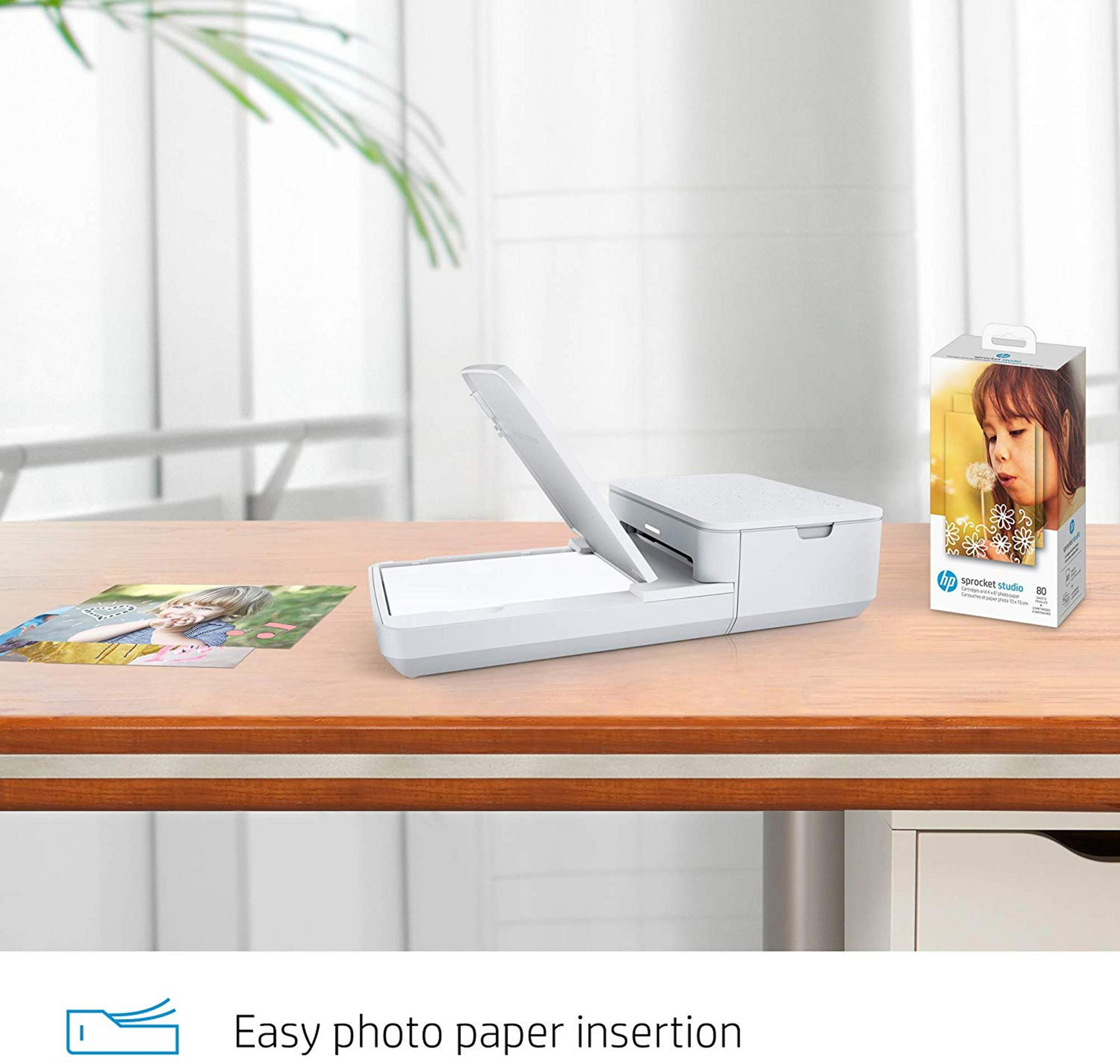 Biała drukarka HP Sprocket Studio z papierem i zdjęciami na drewnianym biurku.