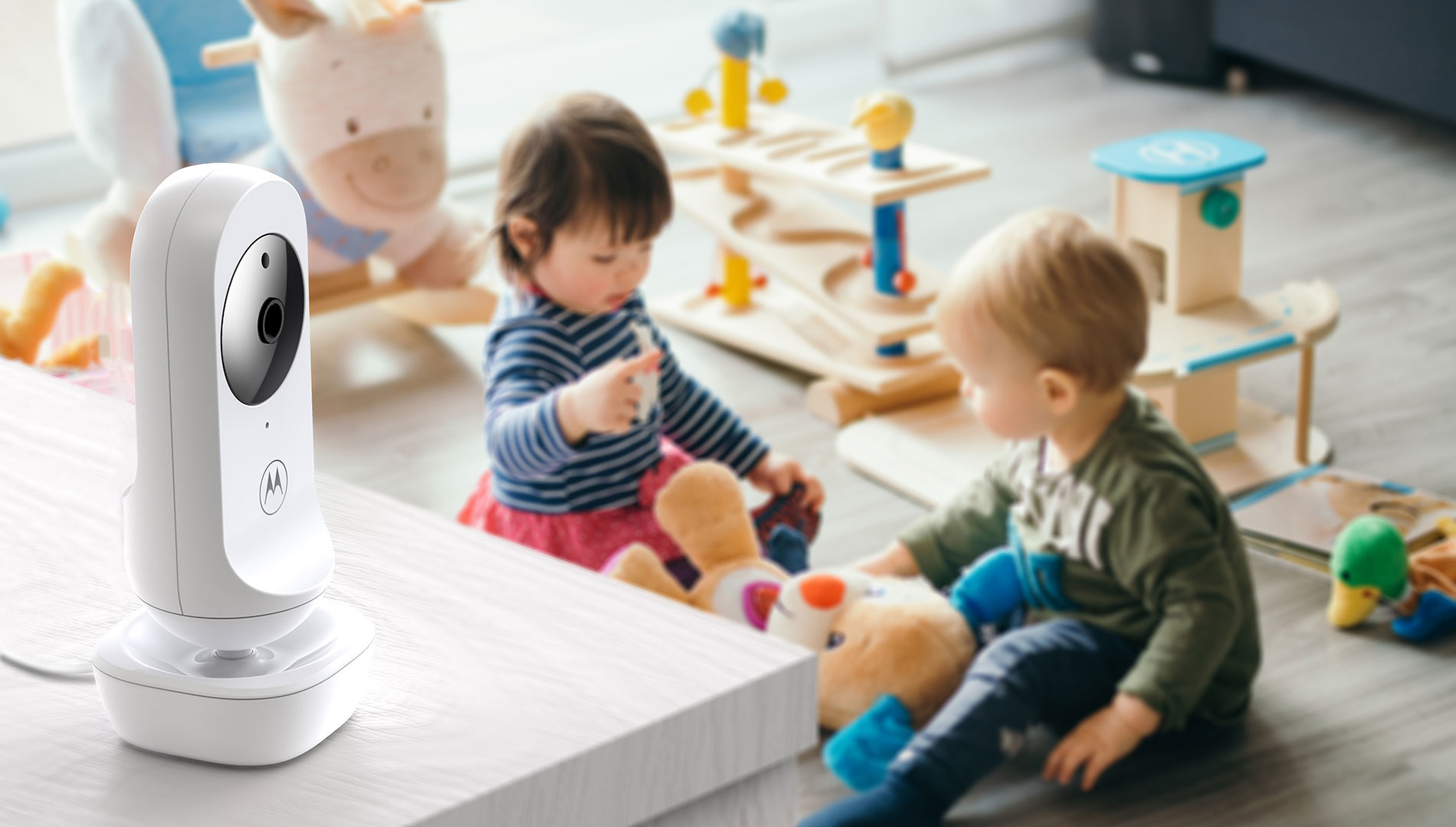Ein weißes Babyphone steht im Vordergrund, zwei Babys spielen mit Spielzeug.