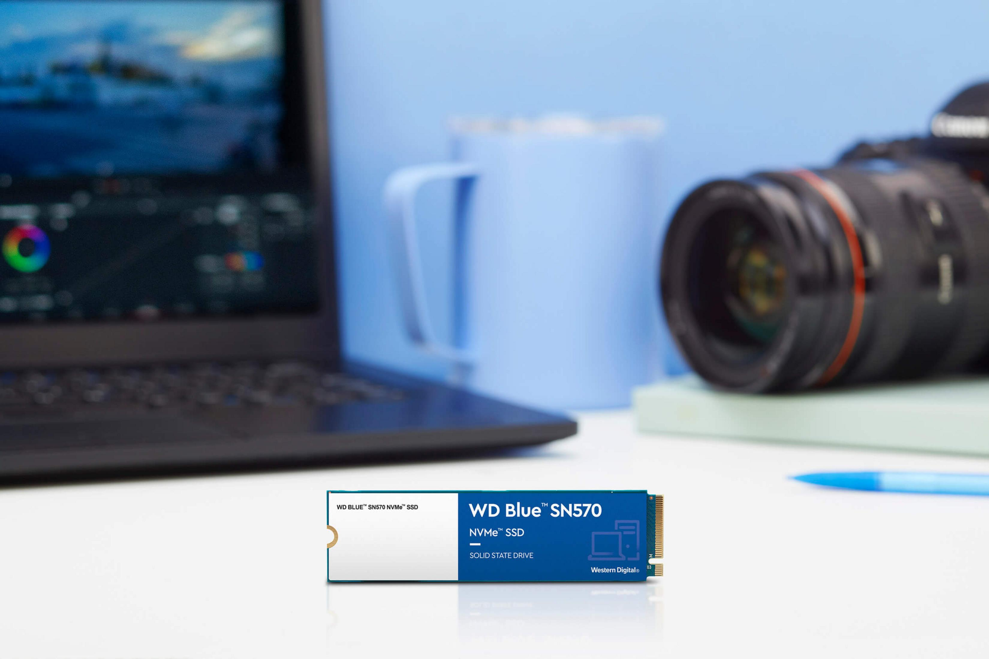Dysk SSD WD Blue SN570 NVMe leży na biurku z laptopem i aparatem.
