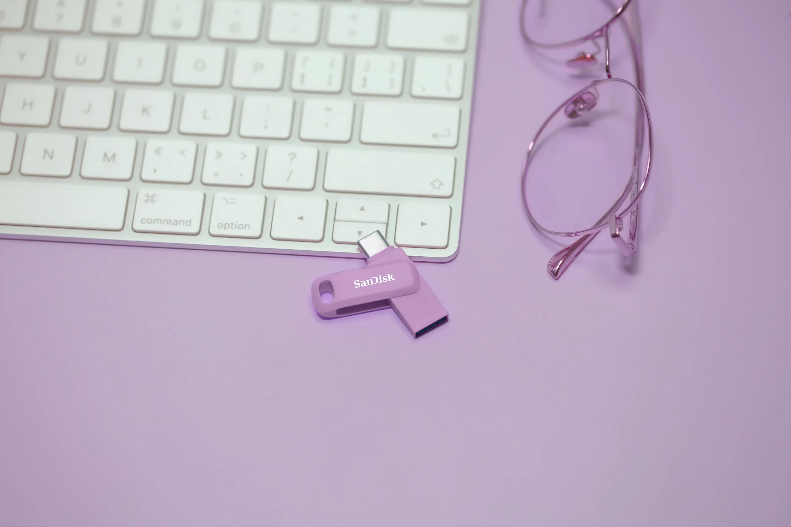Fioletowy pendrive leży obok klawiatury i okularów.