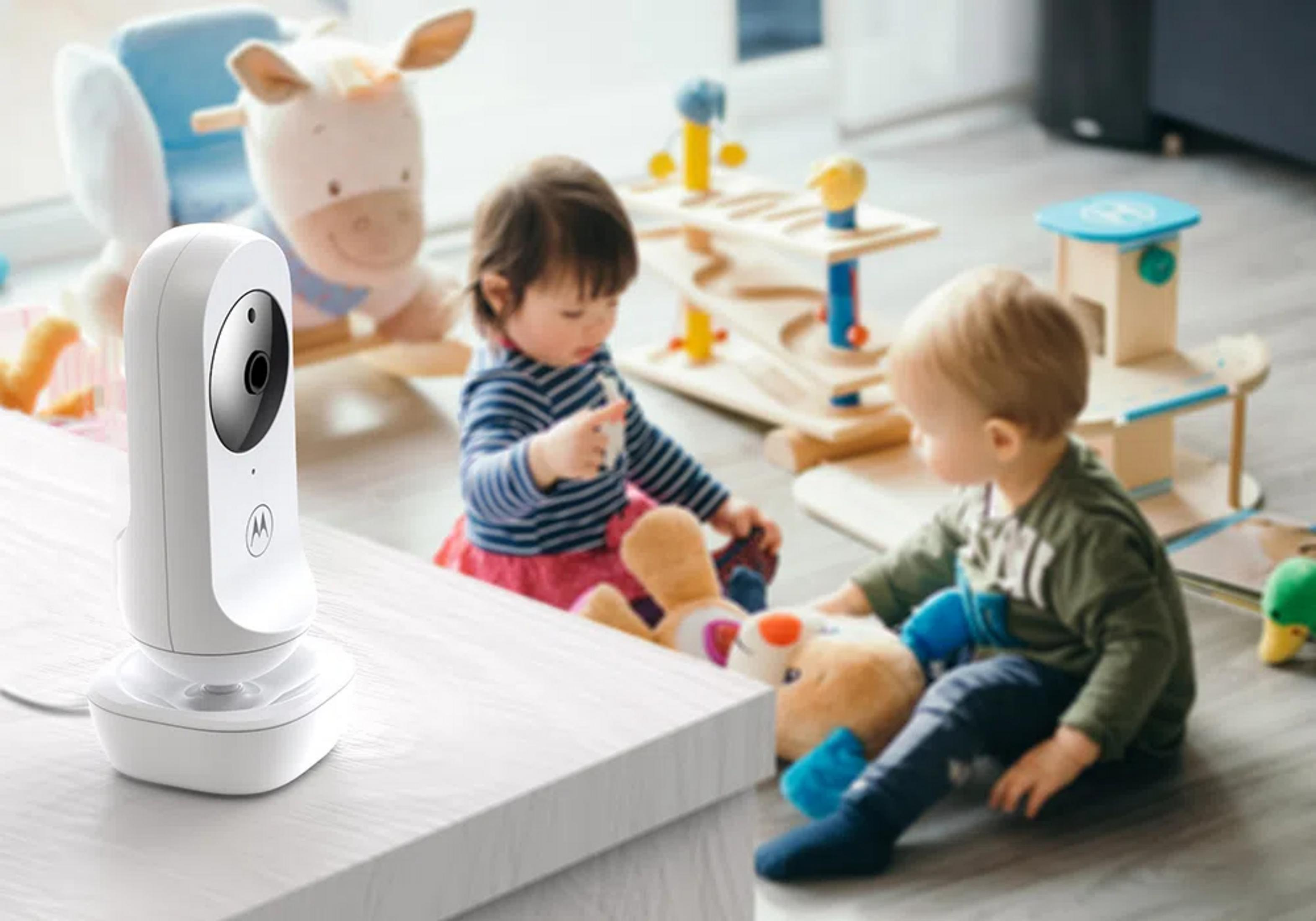 Ein Babyphone steht auf einem Tisch, zwei Kleinkinder spielen mit Spielzeug.