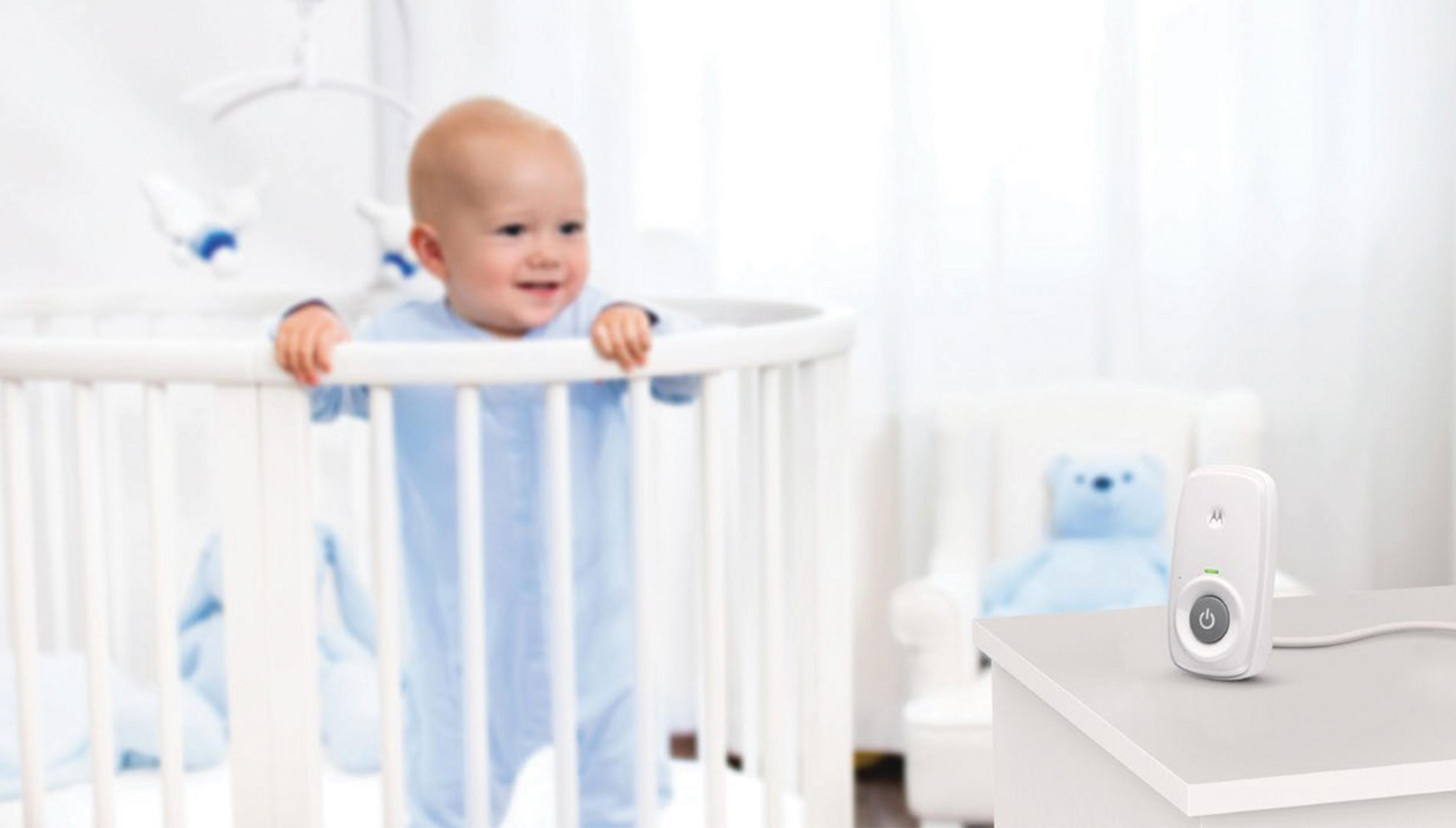 Ein Baby steht in einem Kinderbett, ein Babyfon steht auf einem Tisch daneben.