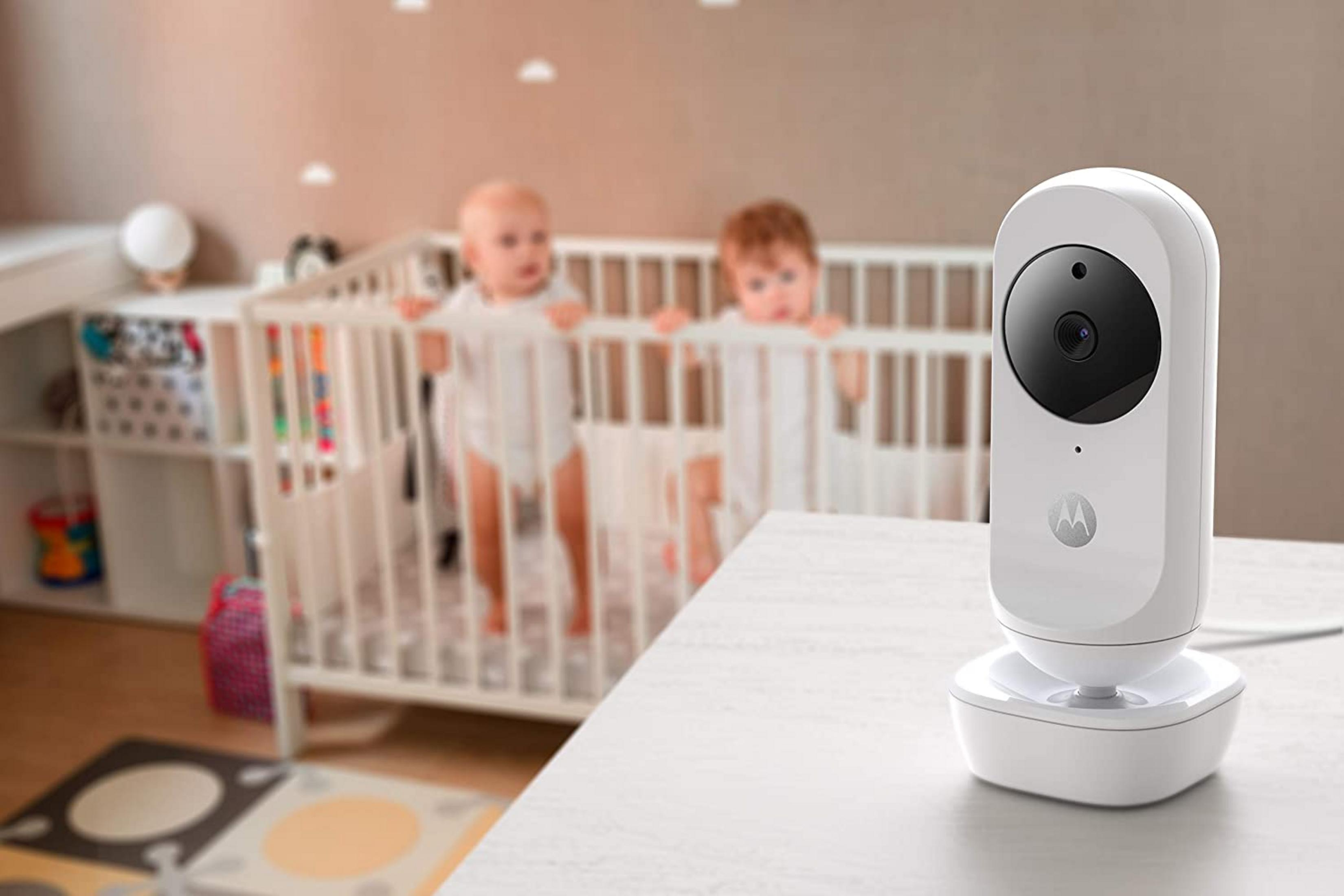 Ein Babyfon steht auf einem Tisch, im Hintergrund zwei Babys im Gitterbett.