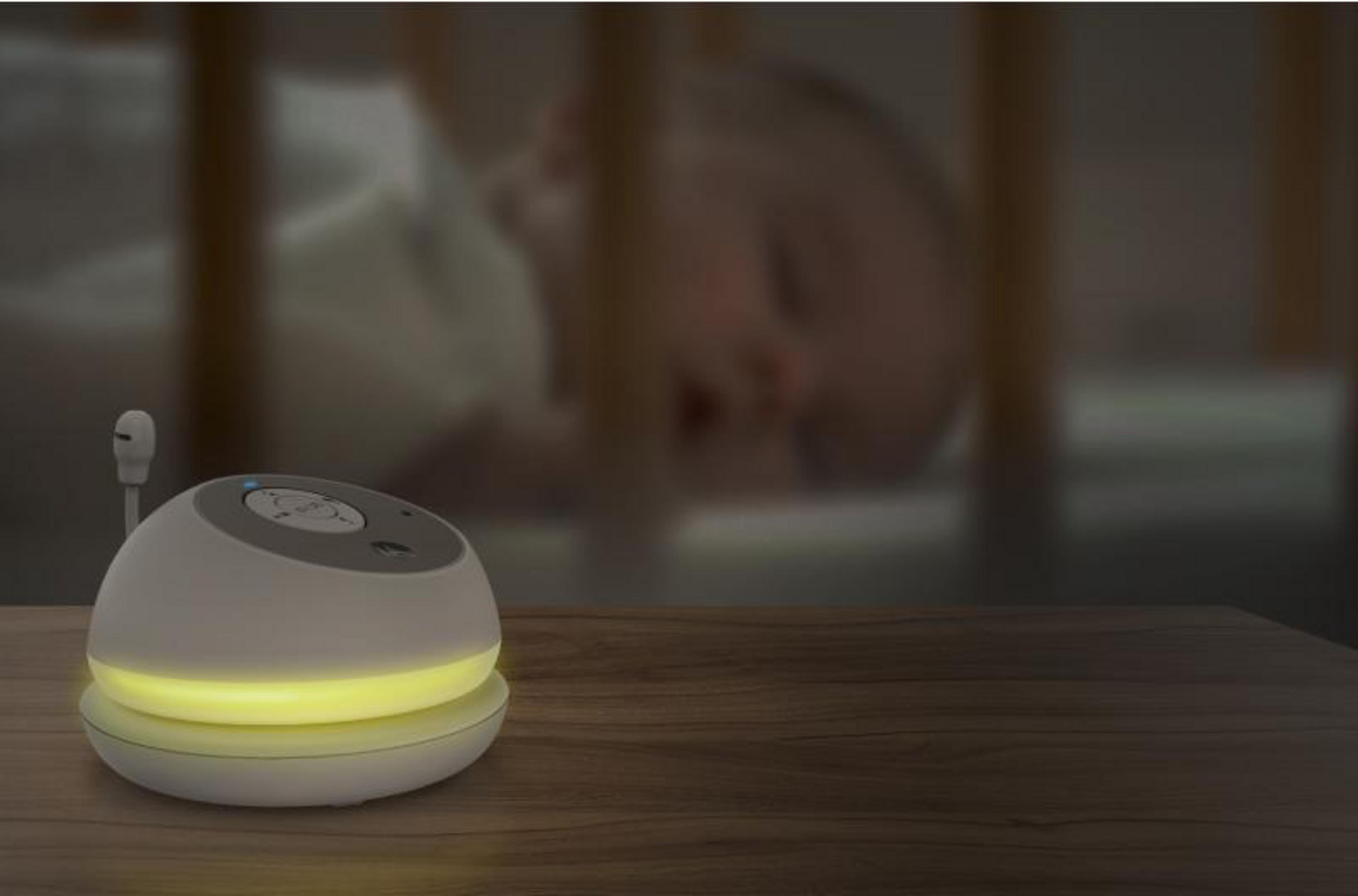 A white baby monitor on a wooden table, a baby sleeps in the background.
