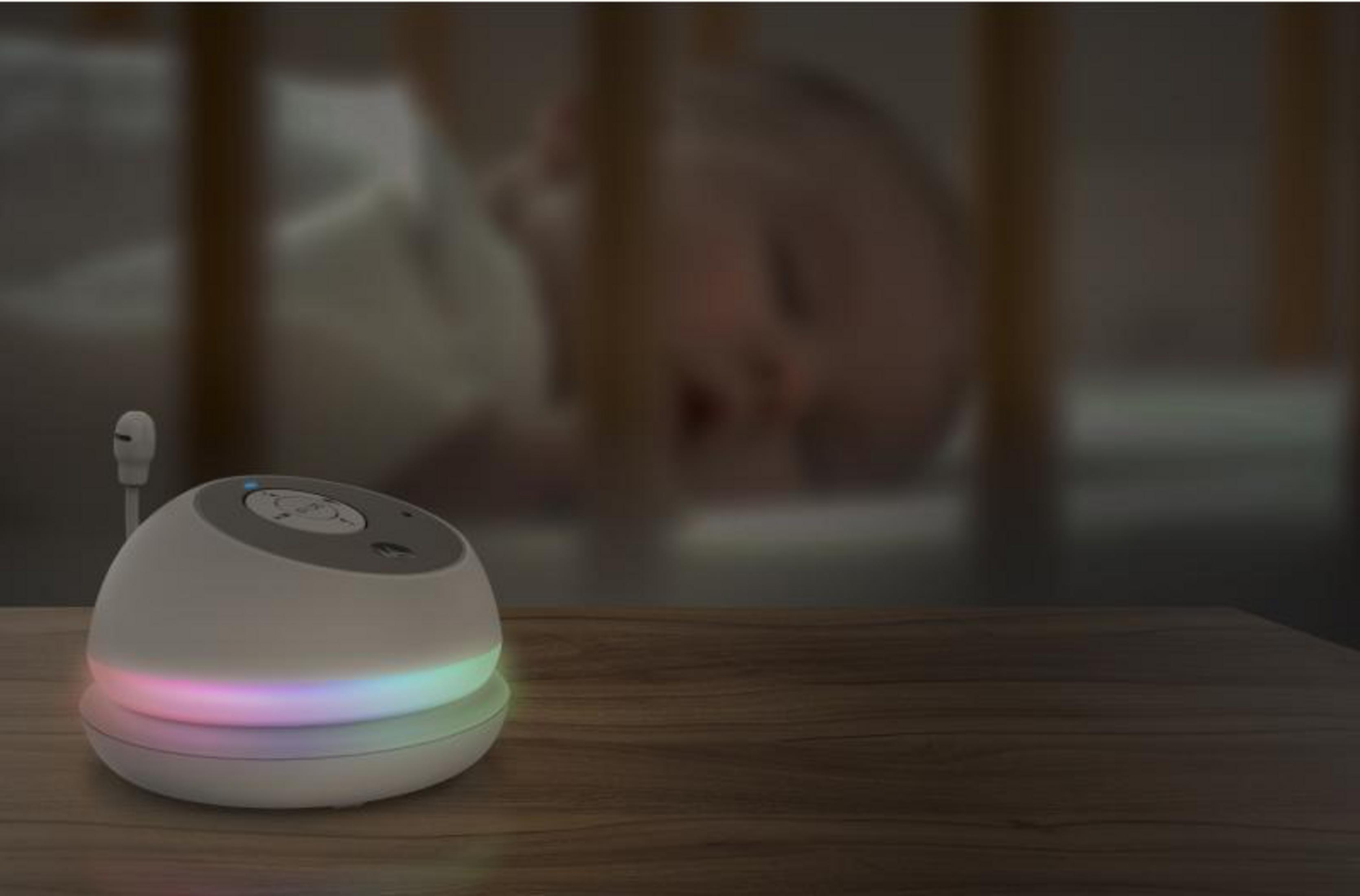 A white baby monitor with colored lights sits on a wooden table. A baby sleeps in a crib in the background.