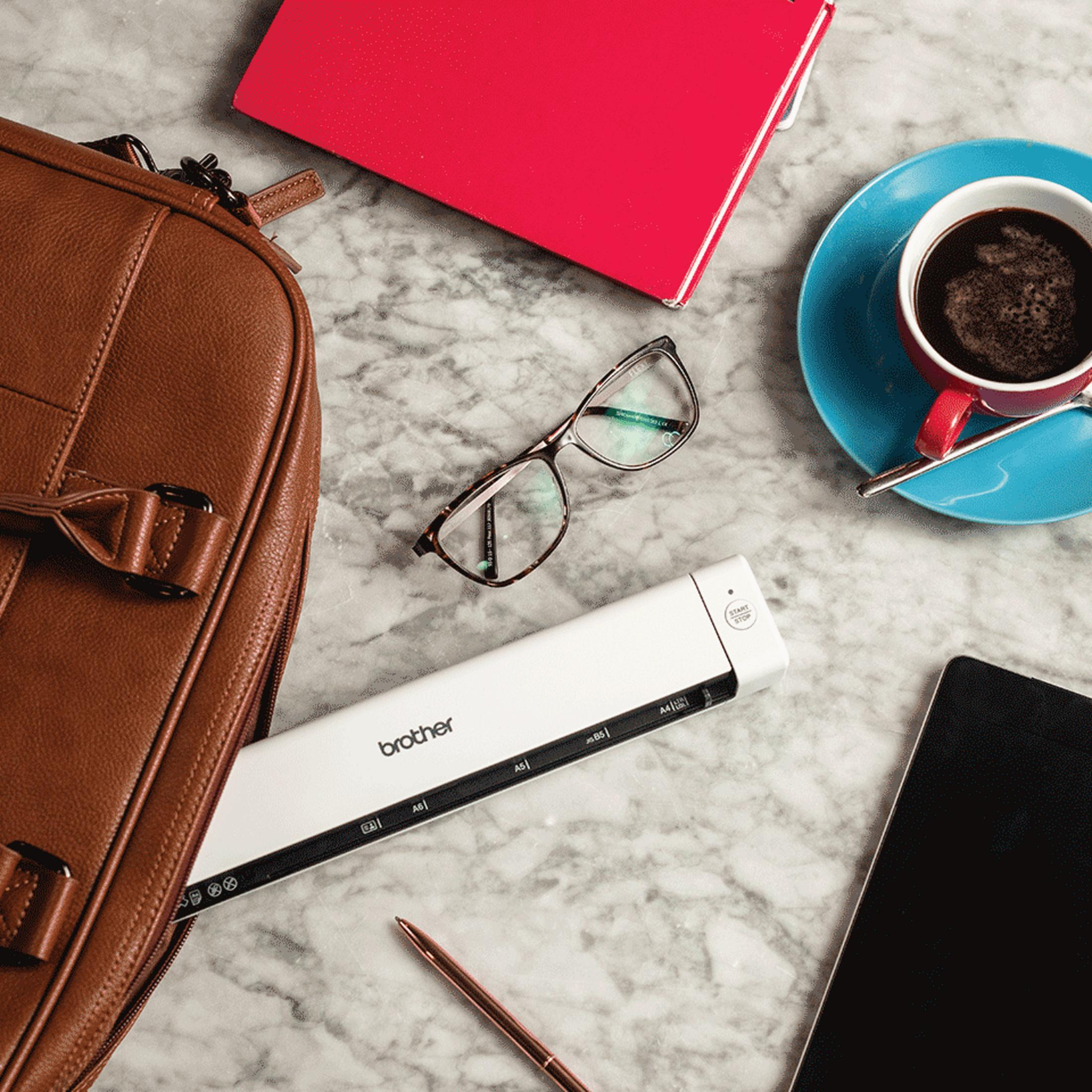 A brown leather bag, red notebook, glasses, scanner, and coffee on a marble surface.