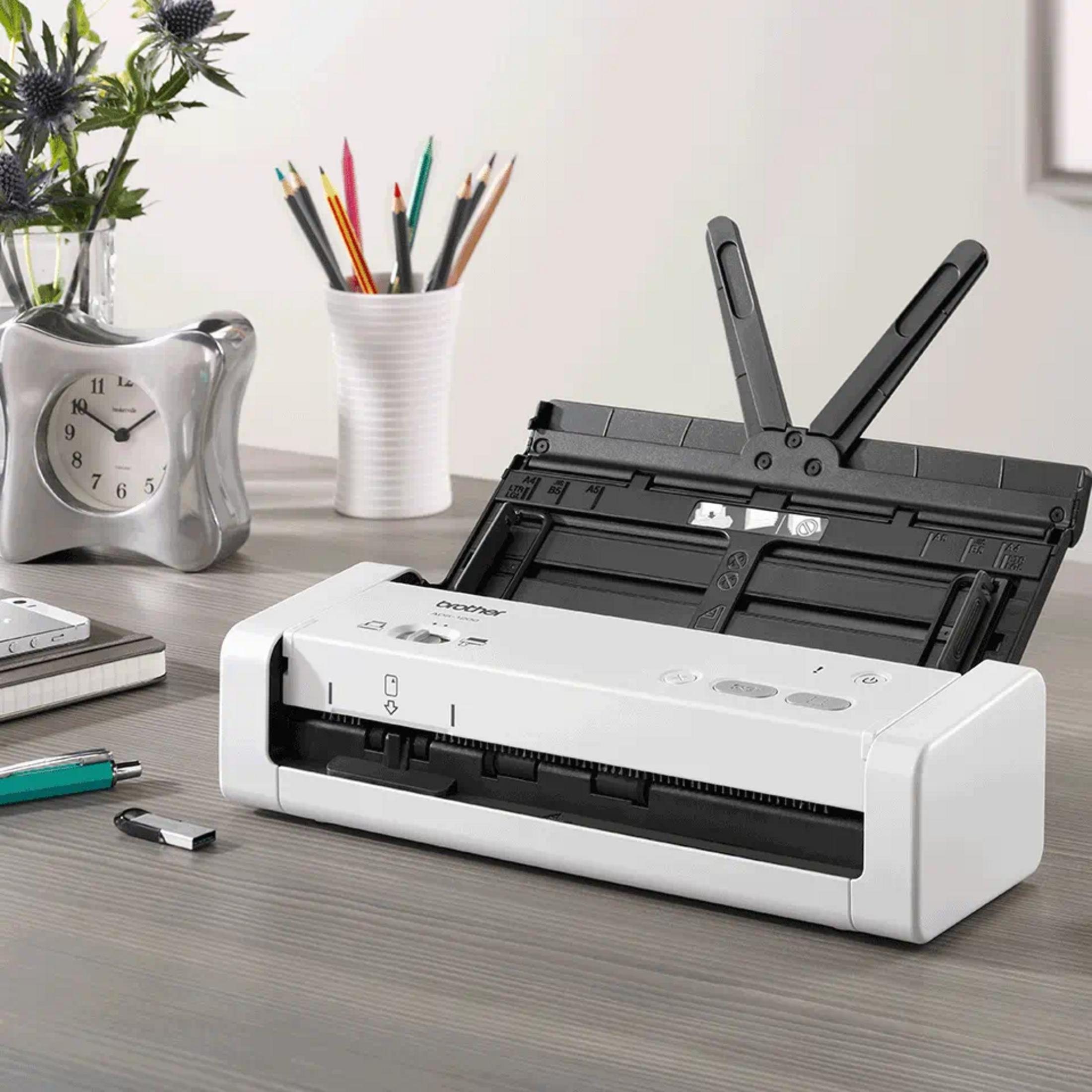 Uno scanner Brother bianco con coperchio aperto si trova su una scrivania di legno accanto a una sveglia argentata.
