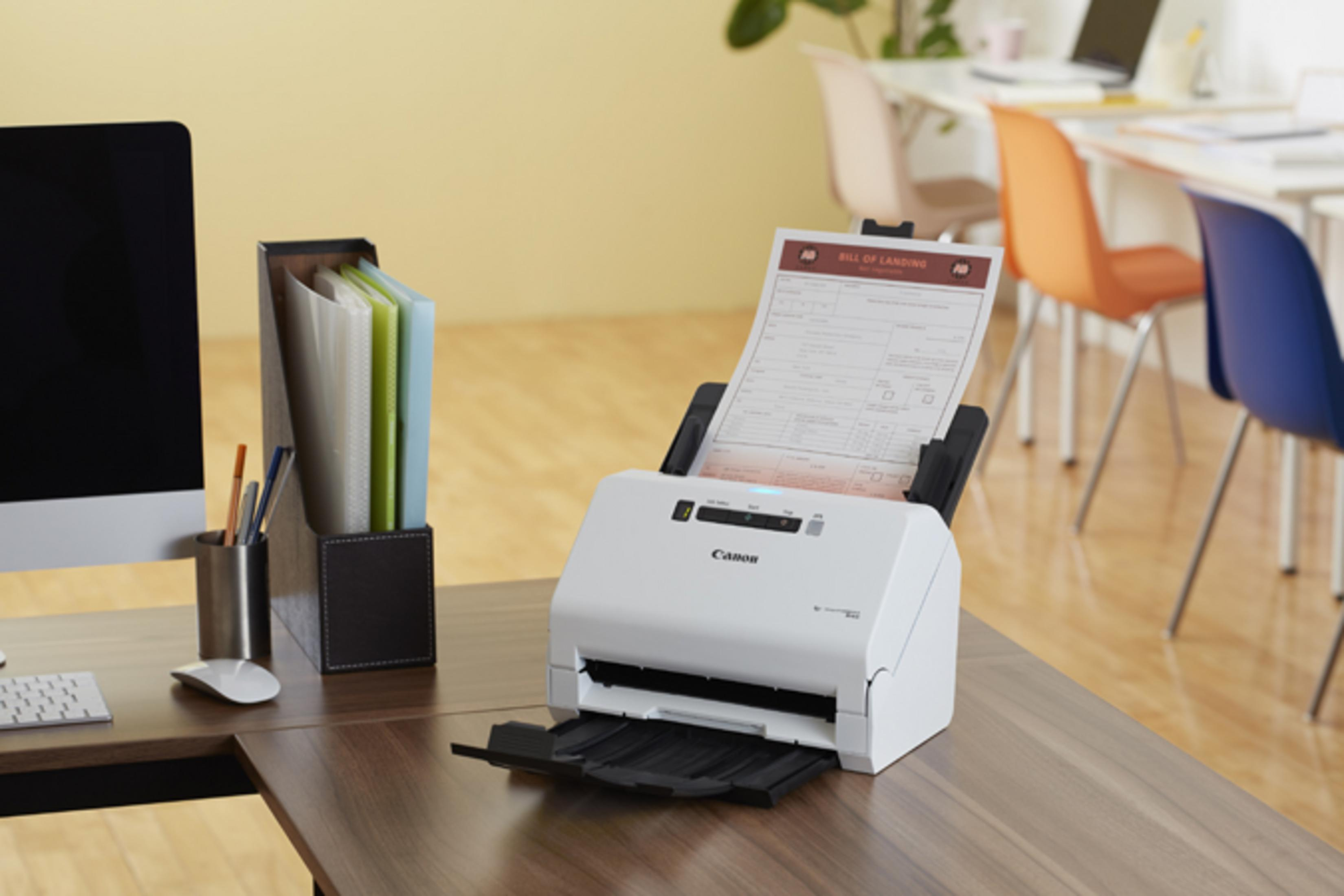 Biały skaner Canon skanuje dokument na biurku. W pobliżu monitor i organizer.
