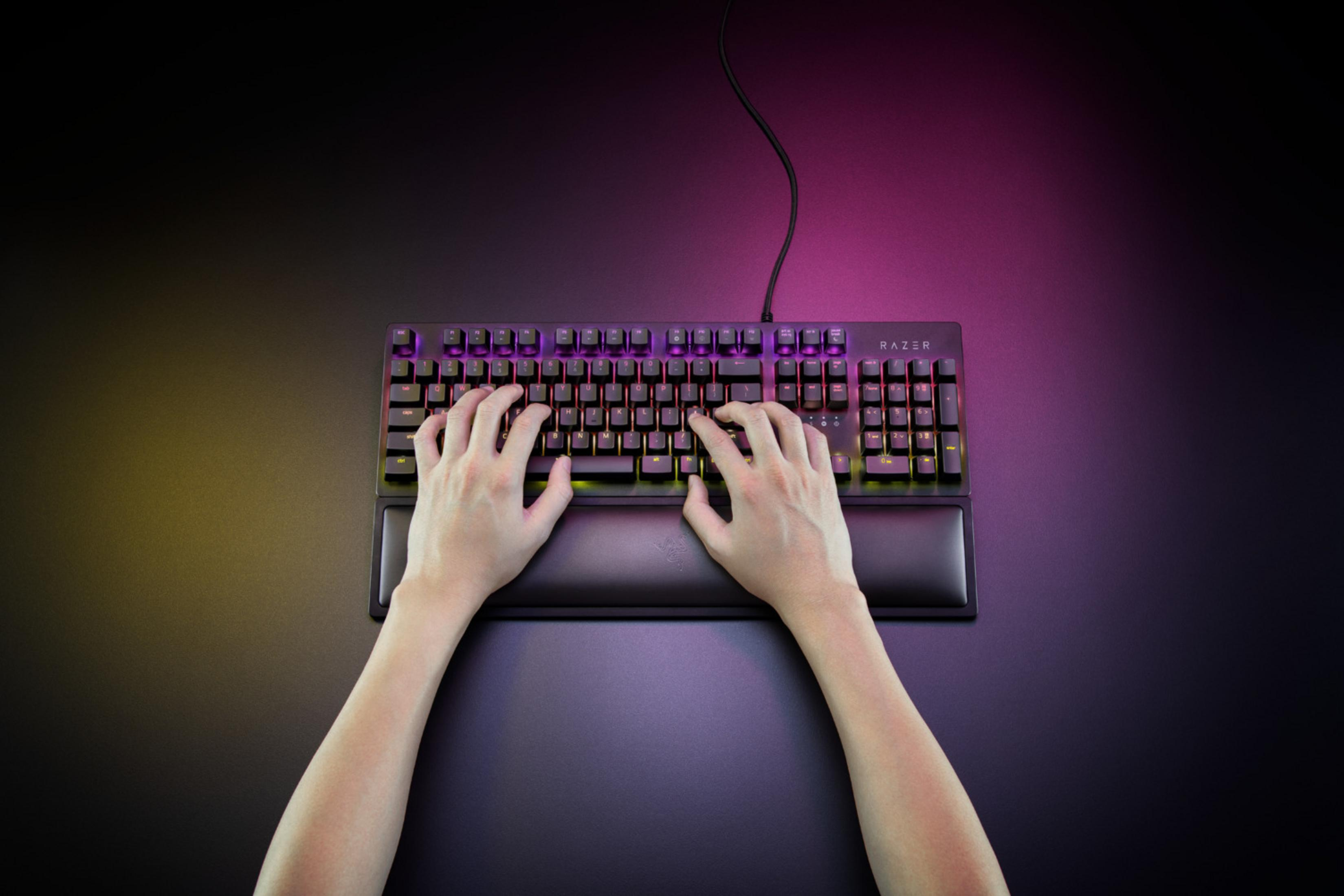 Hände tippen auf einer beleuchteten Tastatur mit Handgelenkauflage, lila und gelbe Beleuchtung.