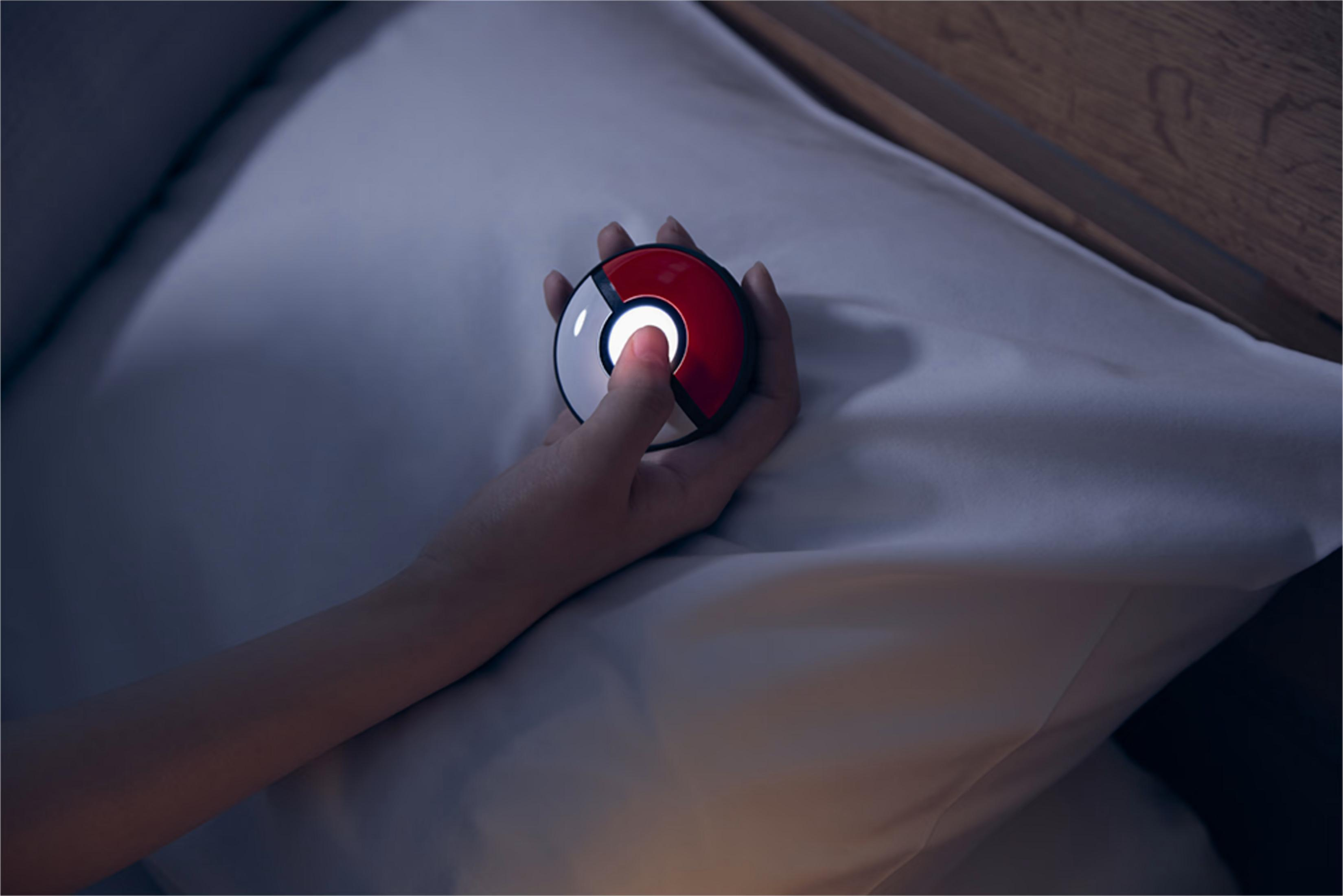 Dłoń trzyma świecący pokeball nad poduszką w łóżku.
