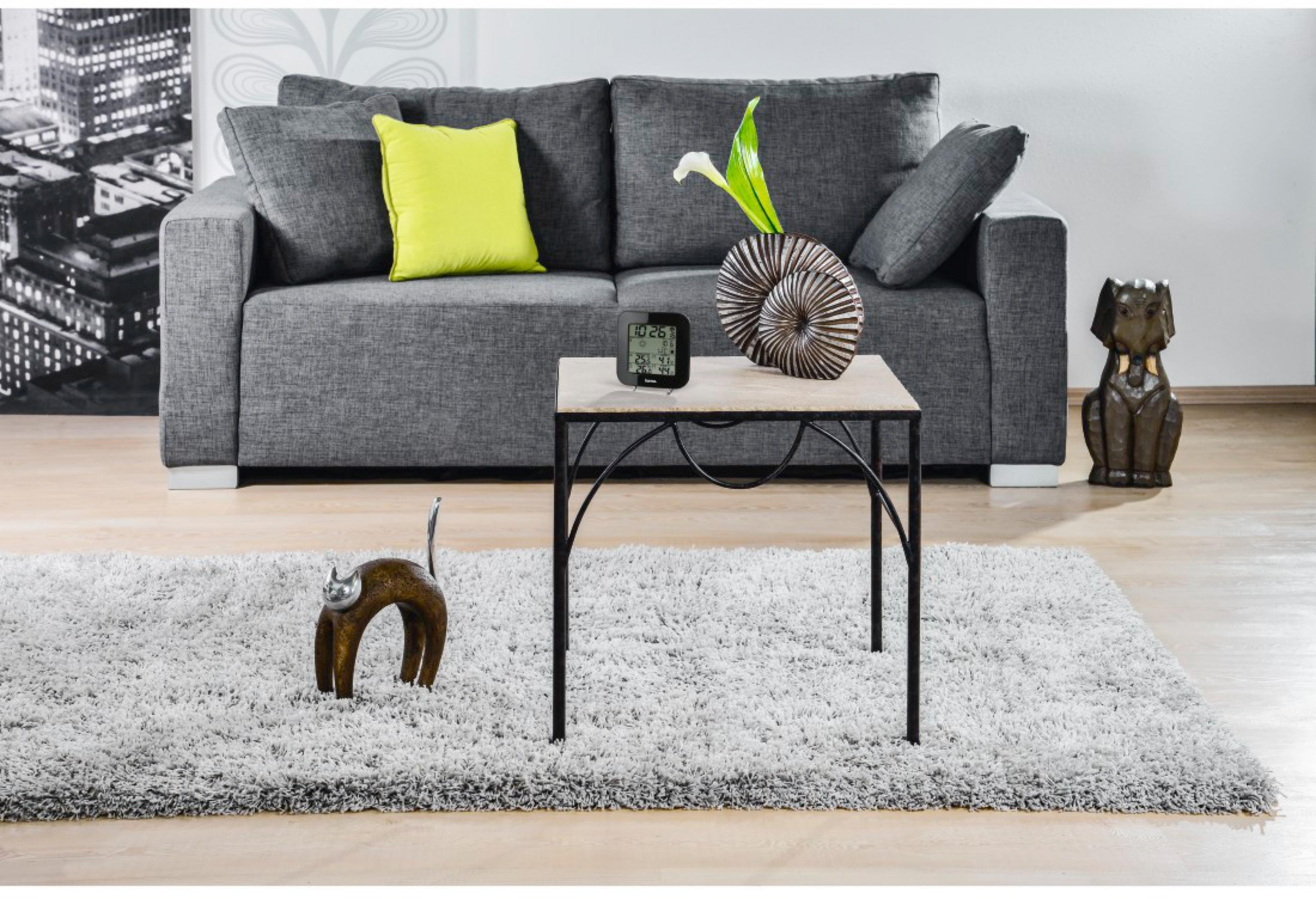 Szara sofa z poduszkami, stolik boczny z dekoracjami i zegarem cyfrowym.