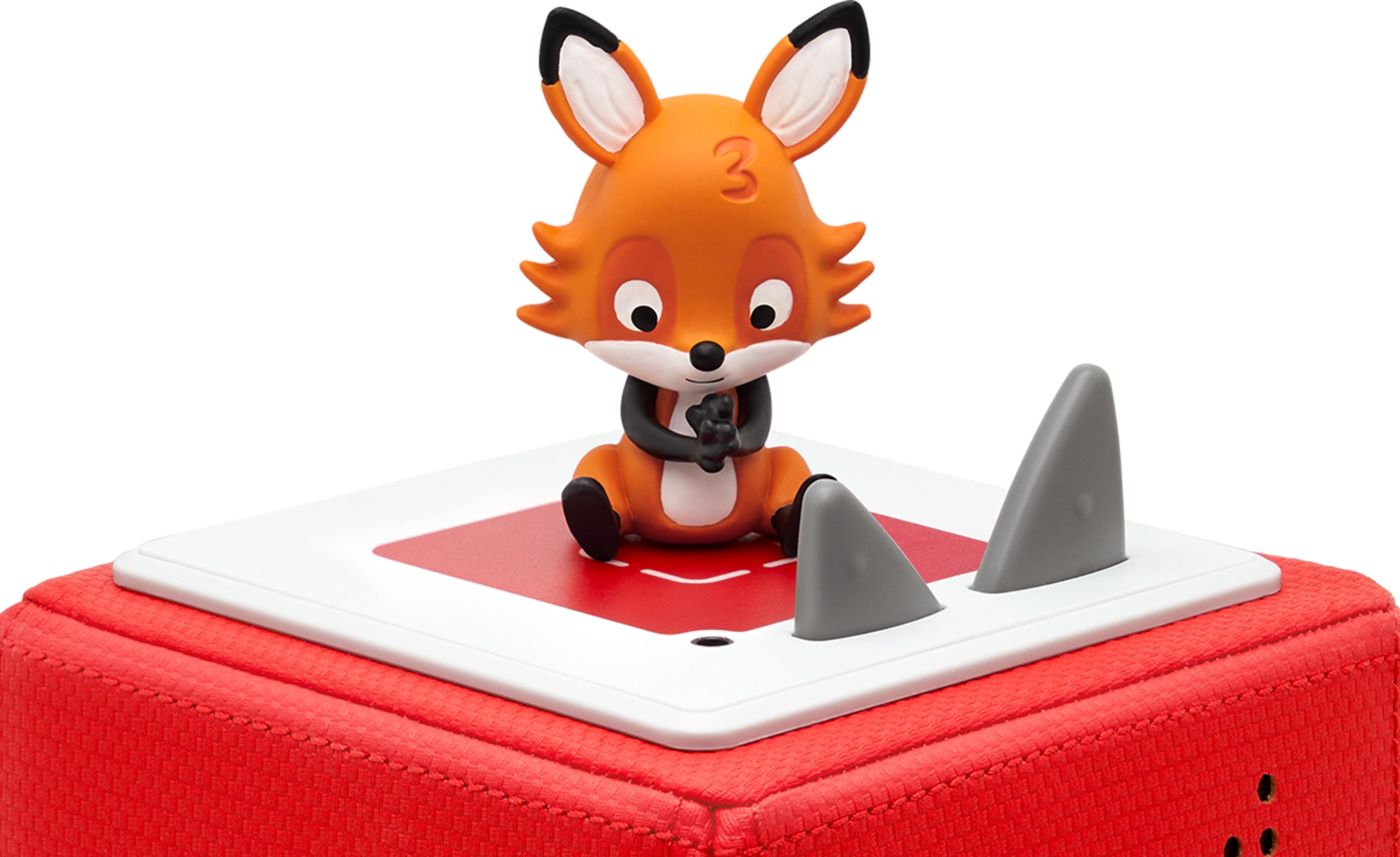 Ein orangefarbener Fuchs sitzt auf einem roten Würfel mit weißen und grauen Elementen.