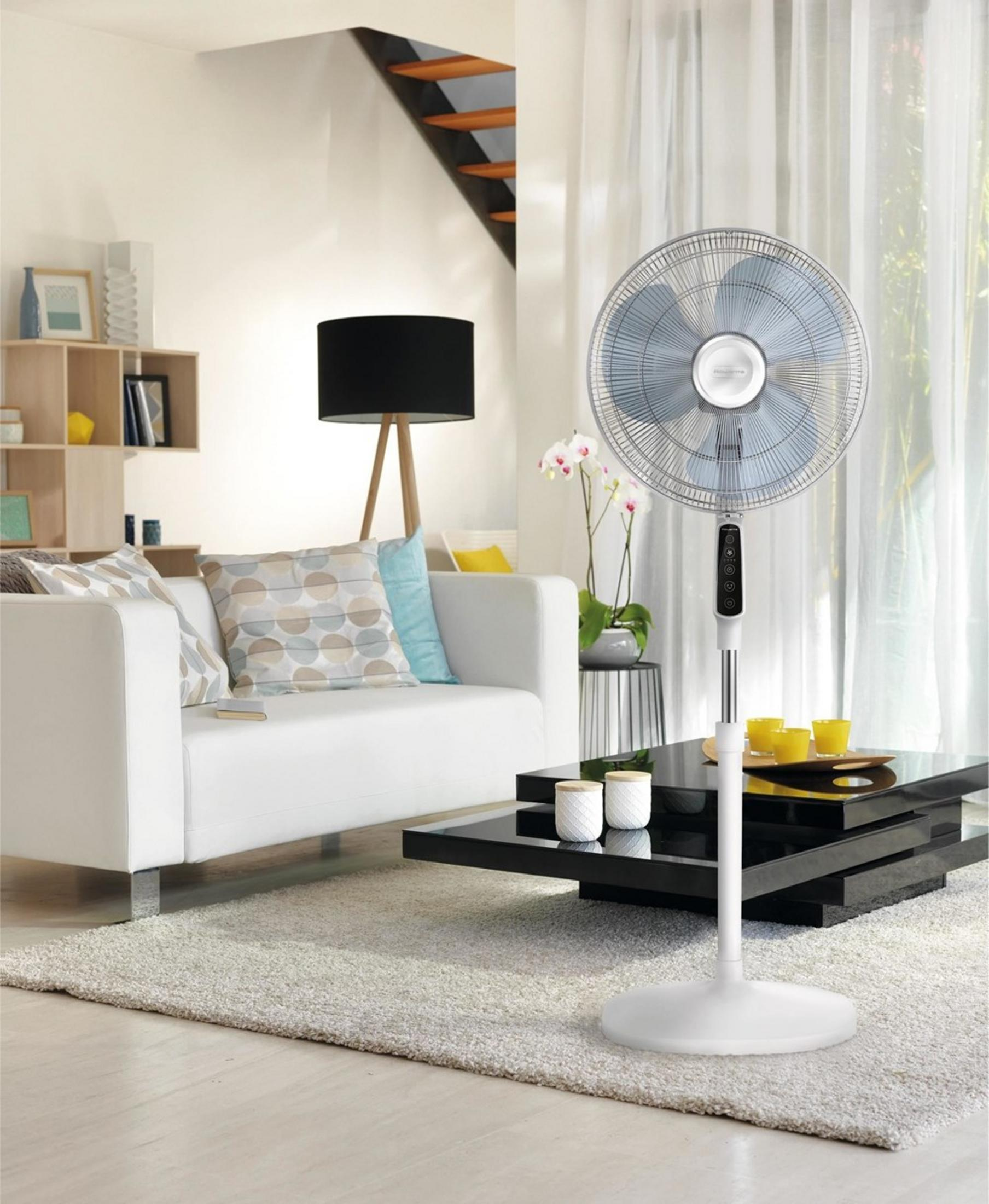 Biały wentylator stojący znajduje się obok białej sofy z poduszkami.
