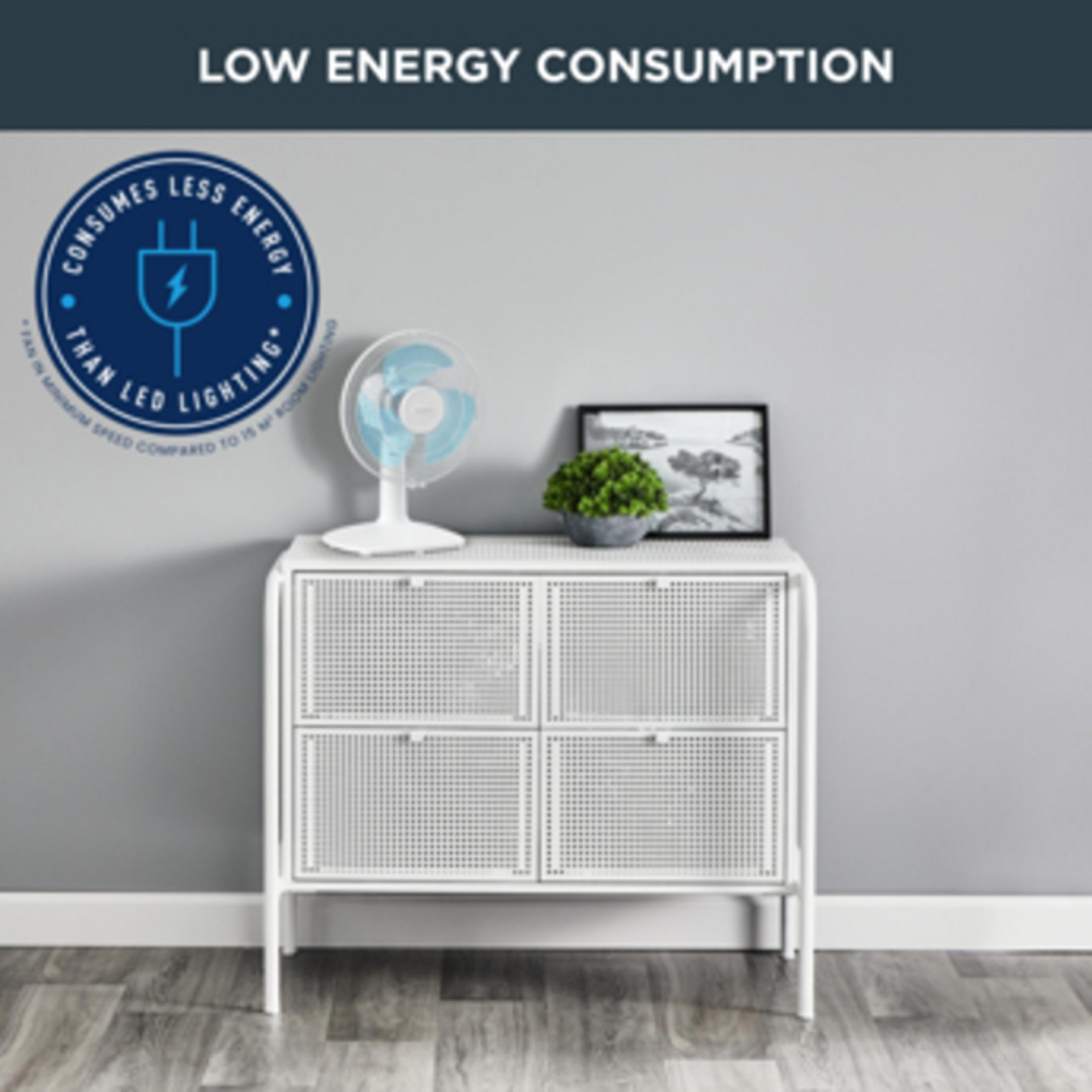 Biały wentylator oscylacyjny na białej szafce z rośliną i oprawioną grafiką.