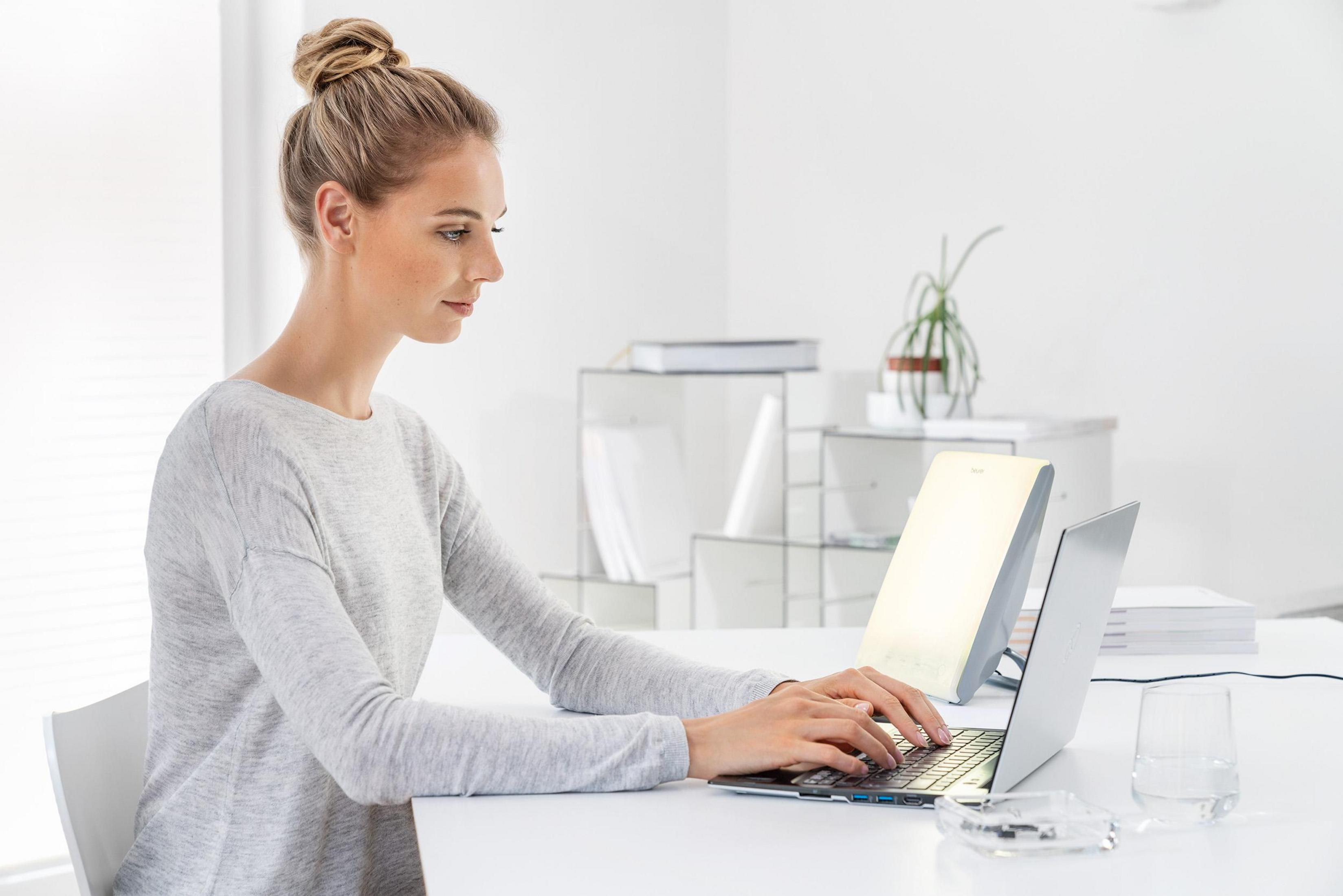 Eine Frau mit Dutt tippt auf einem Laptop, daneben steht eine Lampe.