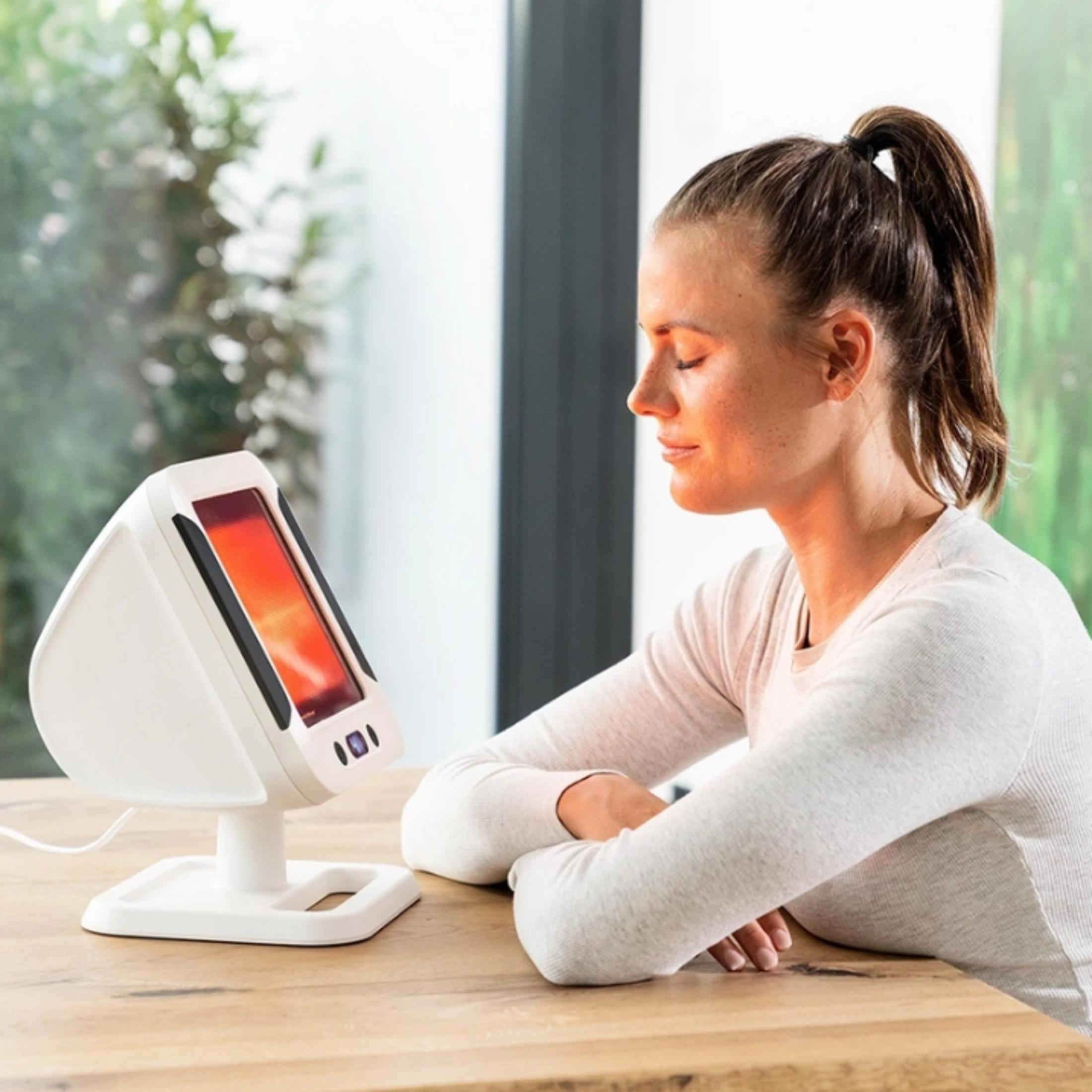 Woman sits with eyes closed in front of a red light therapy device.