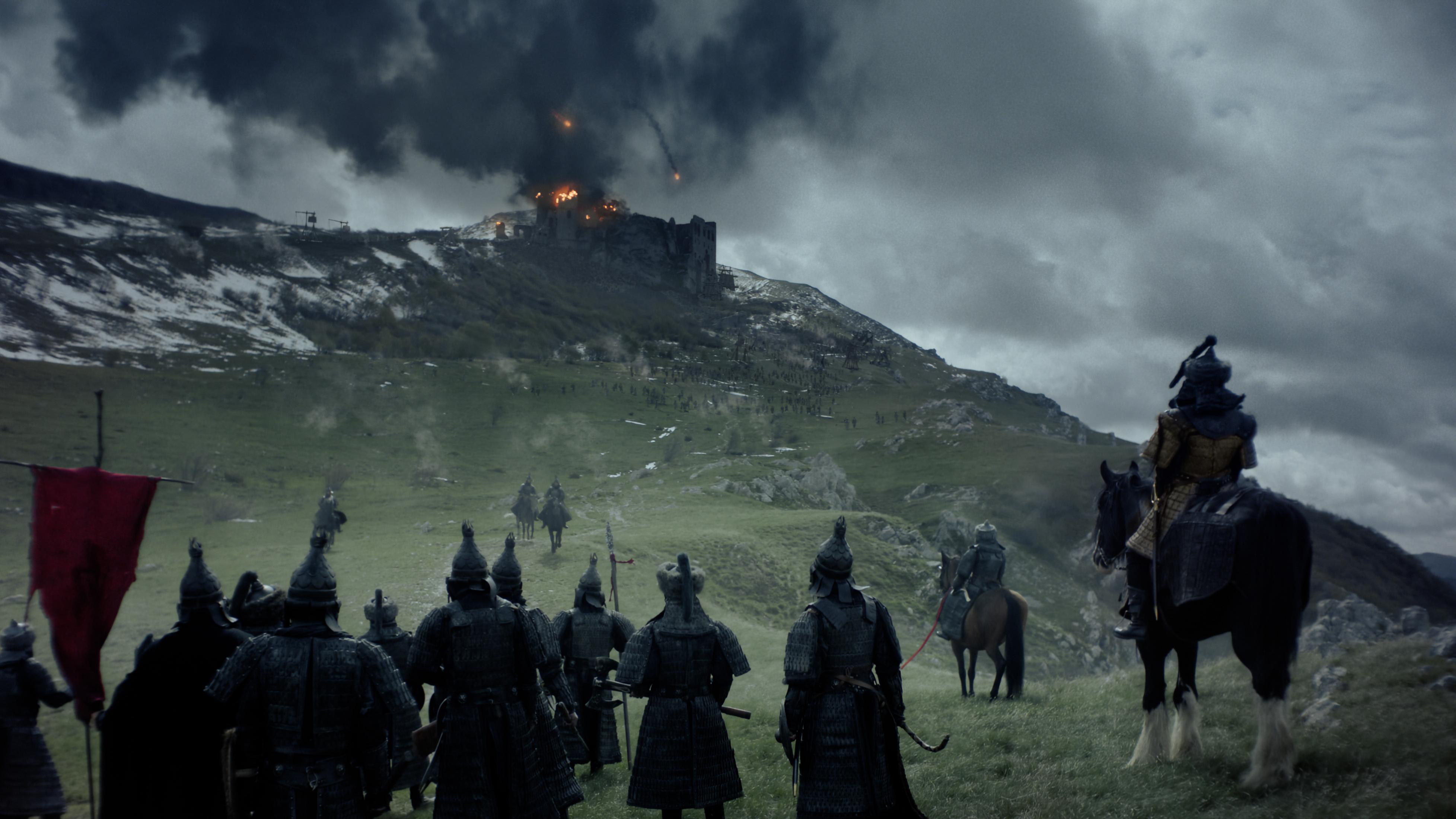 Eine Armee belagert eine brennende Burg auf einem Hügel unter dramatischem Himmel.