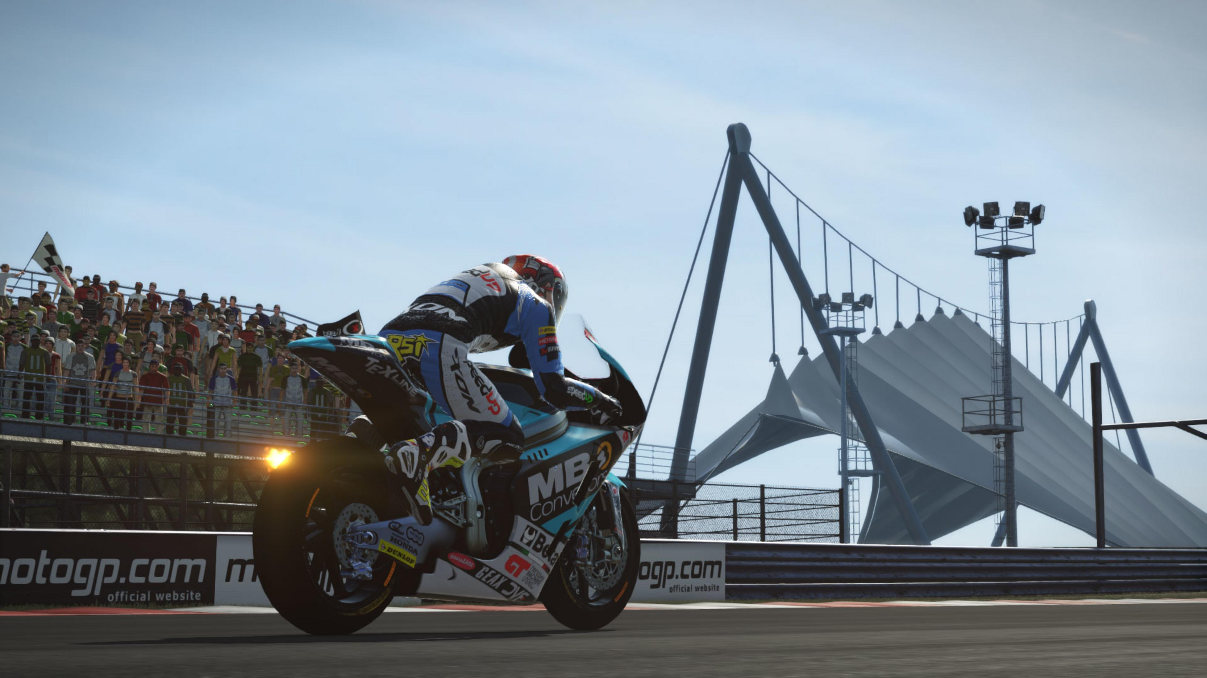 Corridore su moto blu in pista, spettatori sullo sfondo.