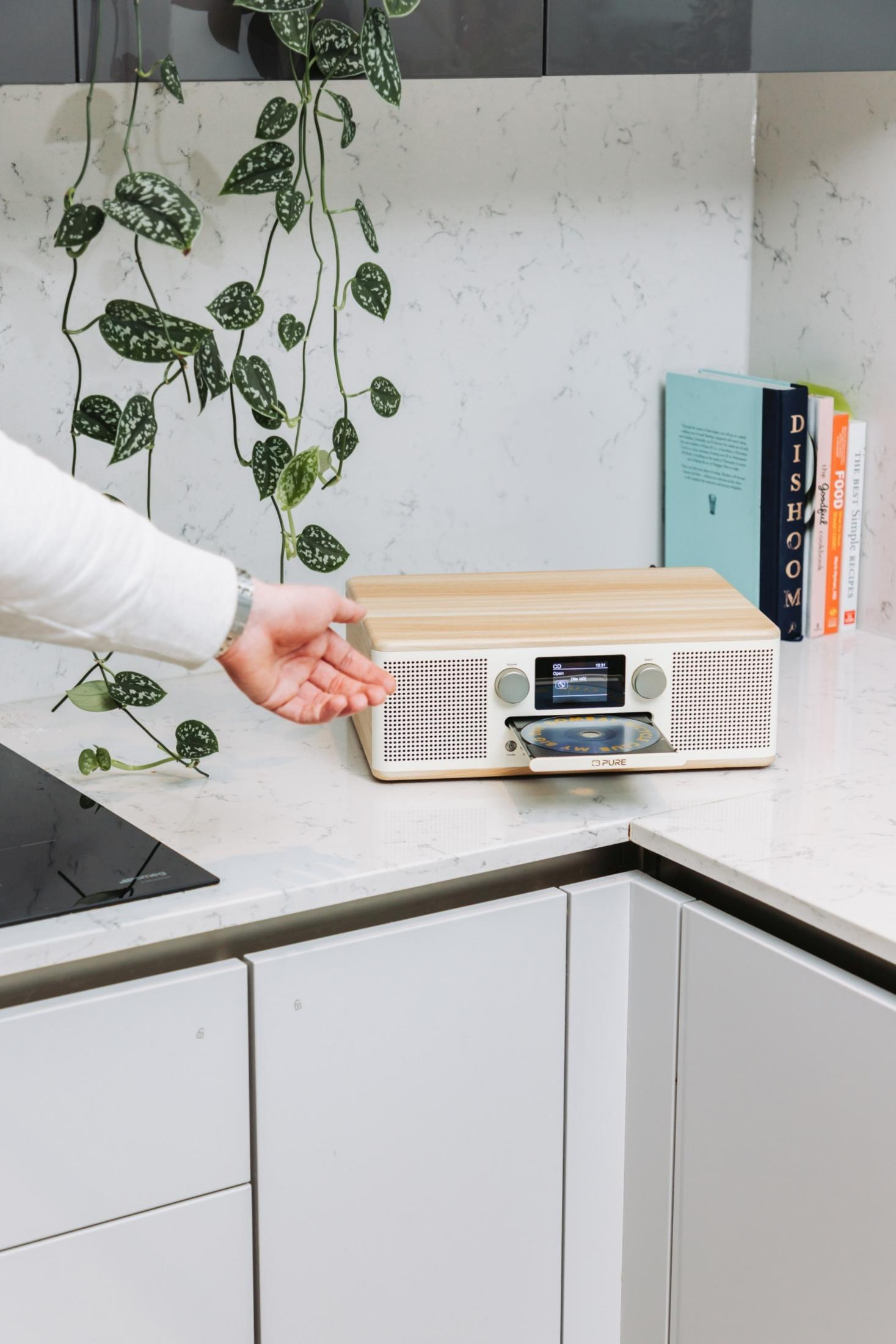 Ręka wkłada płytę CD do jasnego drewnianego radia na marmurowym blacie.