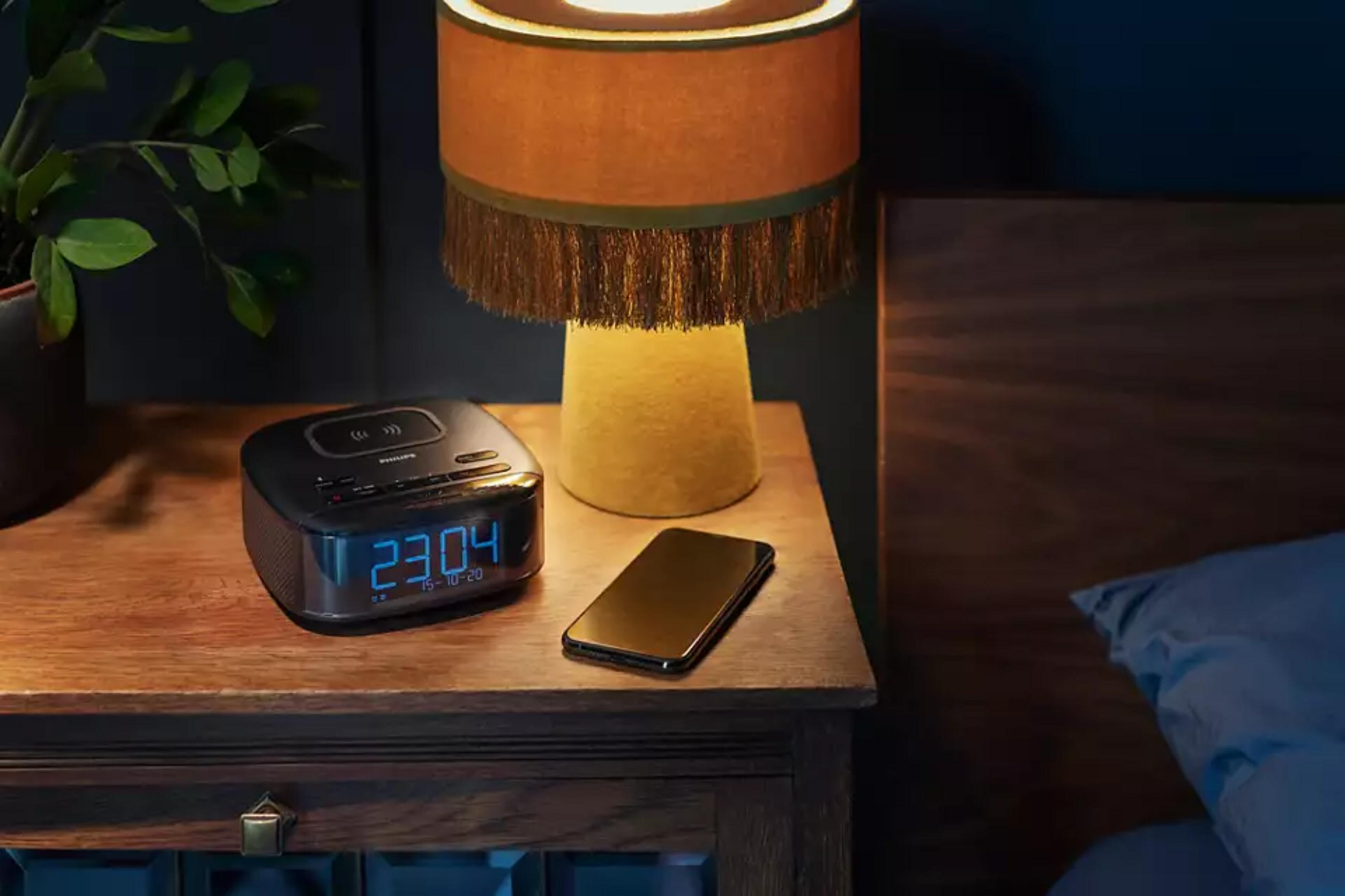 Stolik nocny z zegarem cyfrowym, smartfonem, lampą i rośliną.