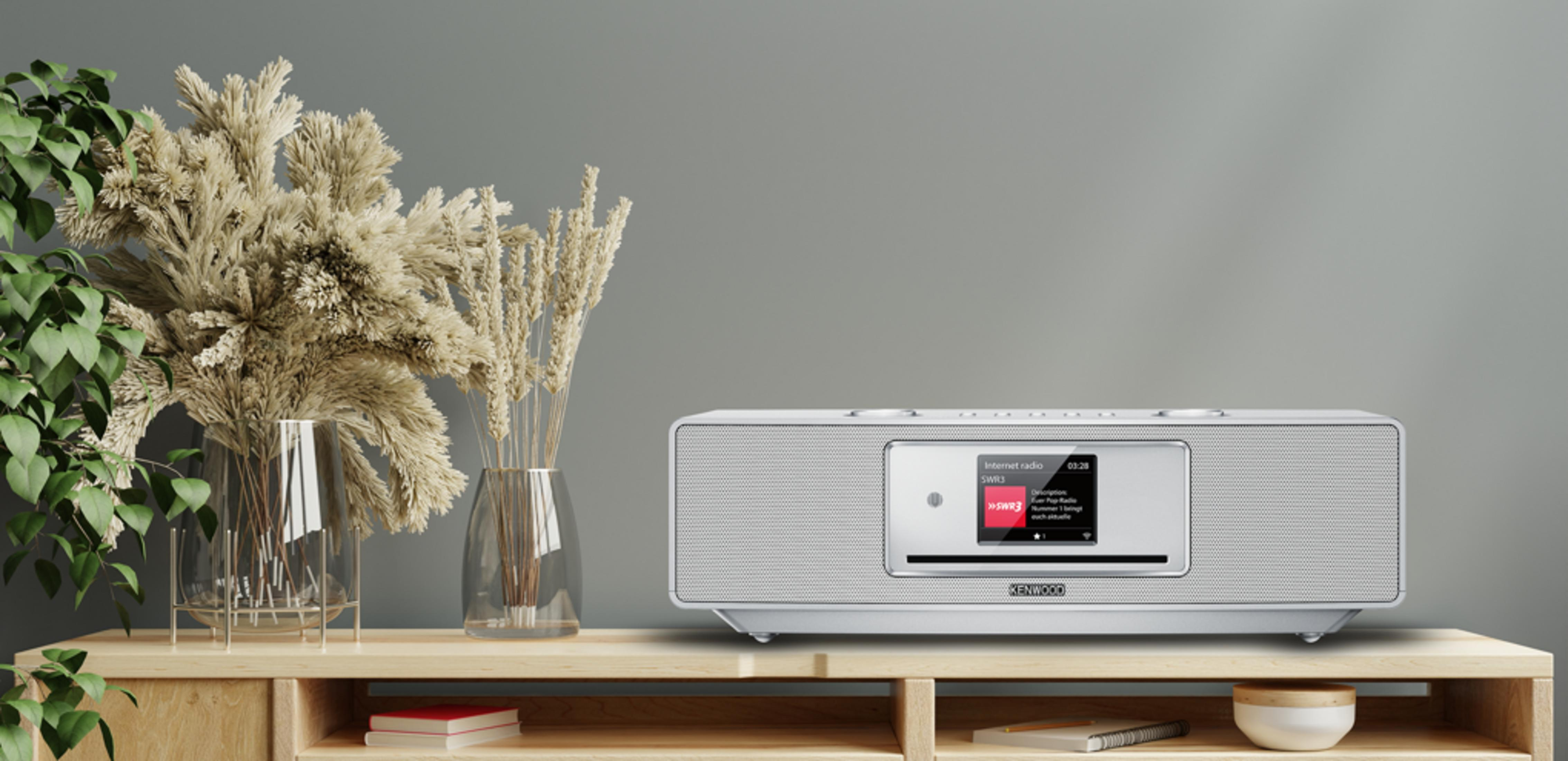 Une radio internet Kenwood blanche sur une étagère en bois à côté d'un vase de fleurs séchées.