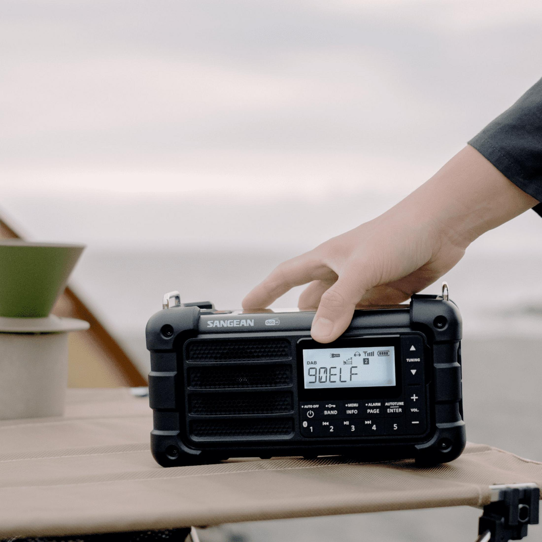 Eine Hand berührt ein schwarzes Sangean DAB+-Radio auf einem Tisch im Freien.