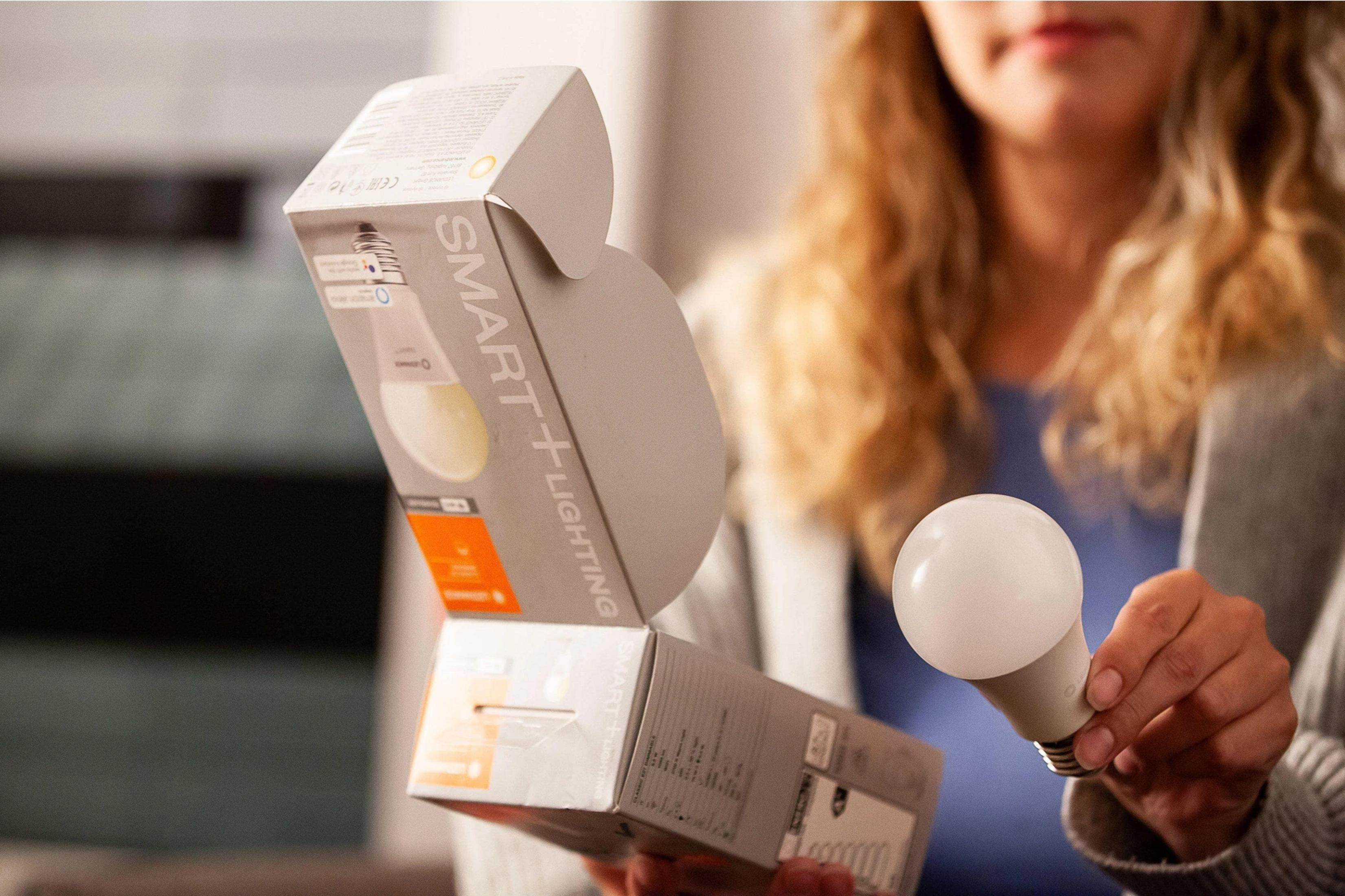 Kobieta trzyma białą żarówkę i otwarte pudełko inteligentnego oświetlenia.