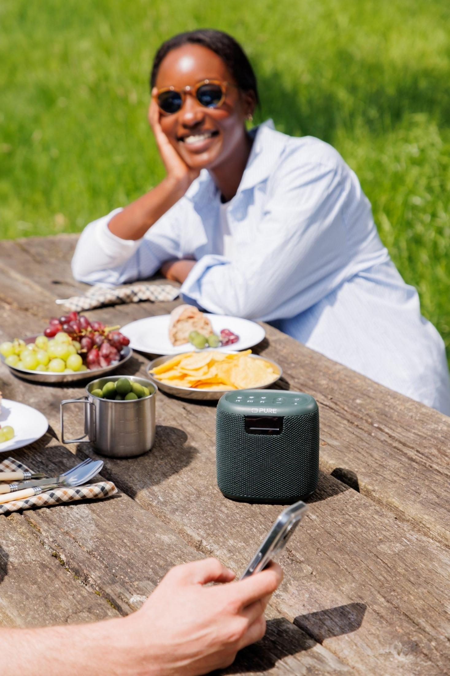 Femme souriante en plein air, pique-nique sur table en bois avec fruits, chips et enceinte.