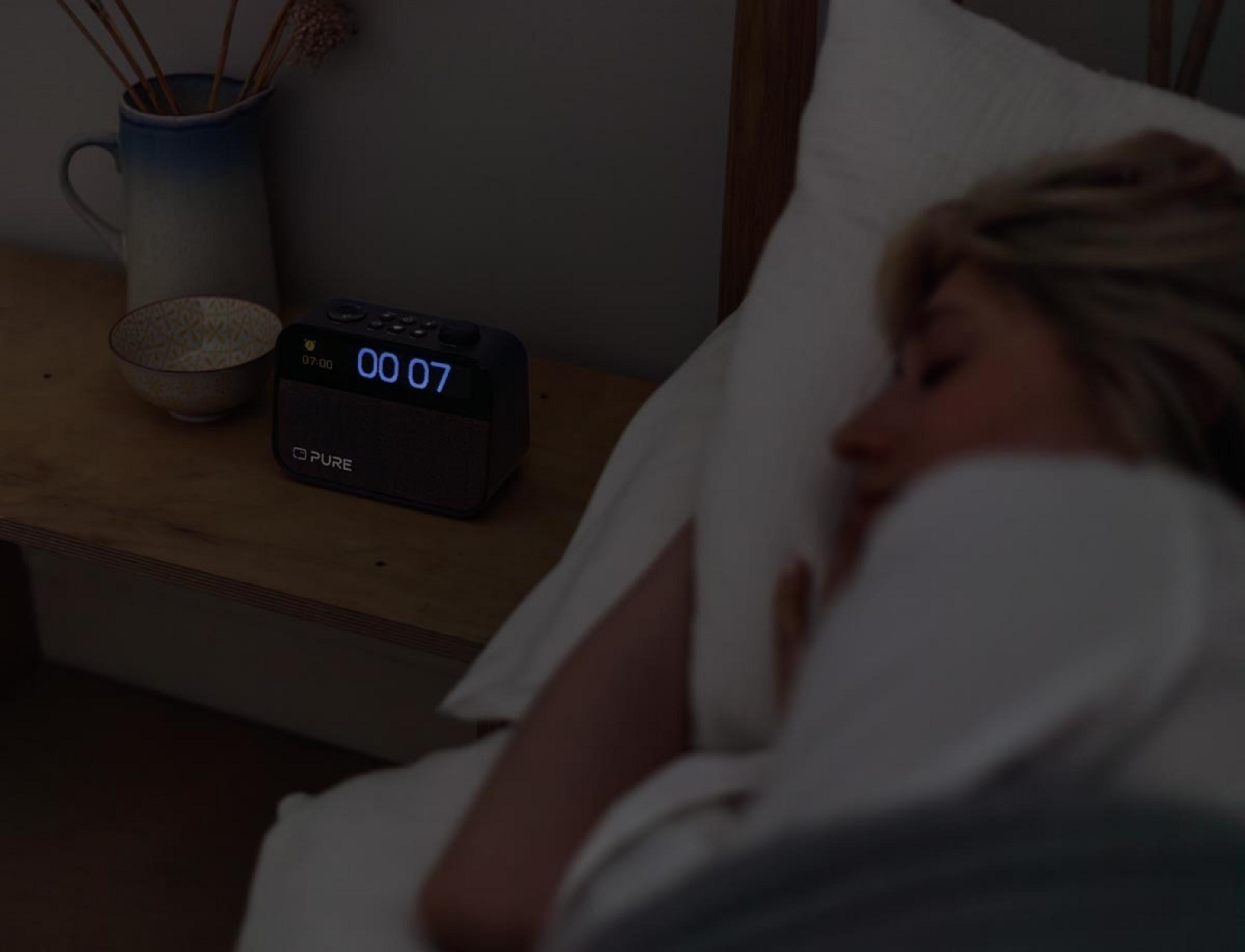 Una mujer duerme en la cama, un reloj digital muestra las 00:07.