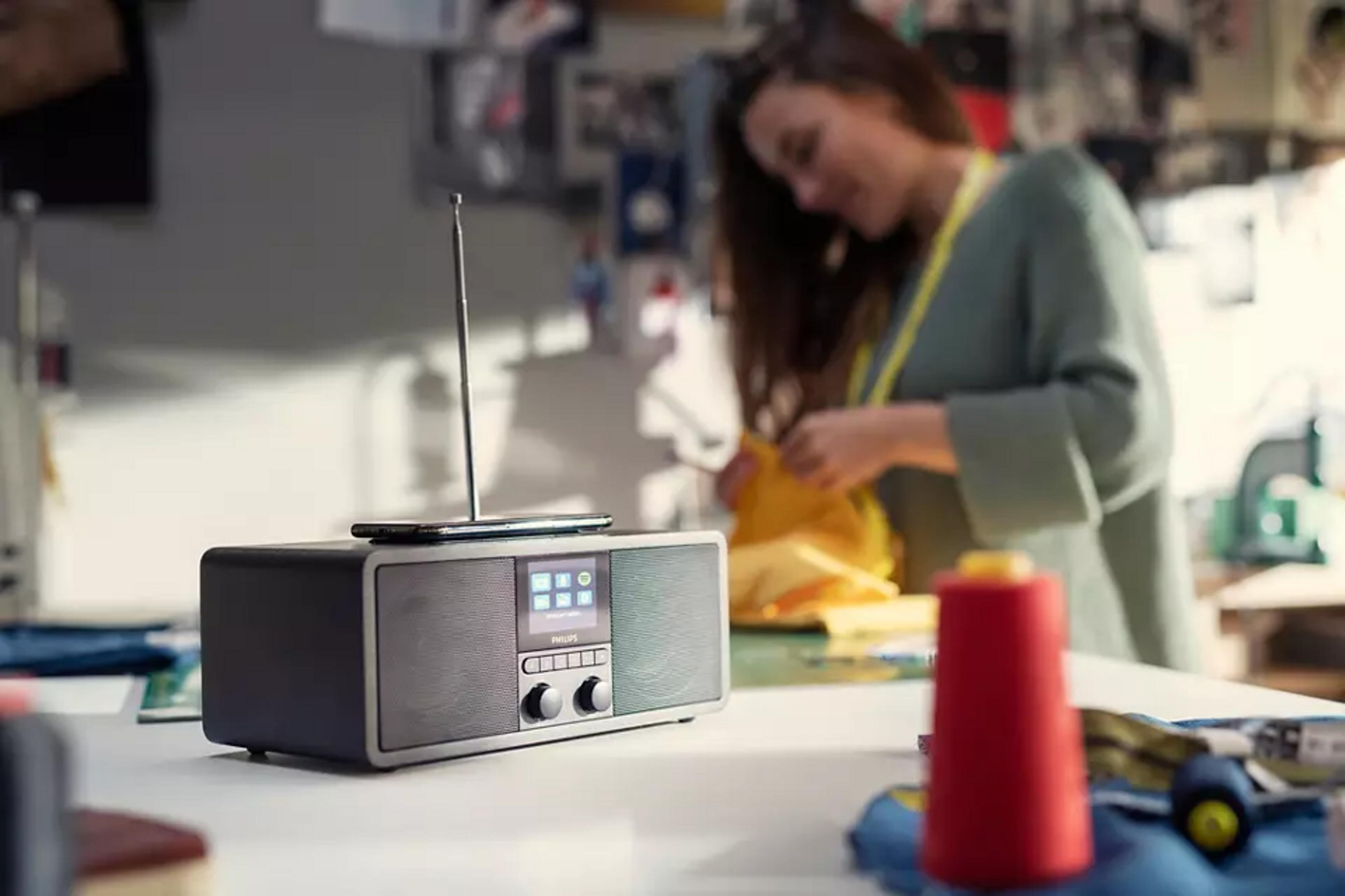 Ein Radio mit Display und Antenne steht auf einem Tisch. Eine Frau arbeitet im Hintergrund.