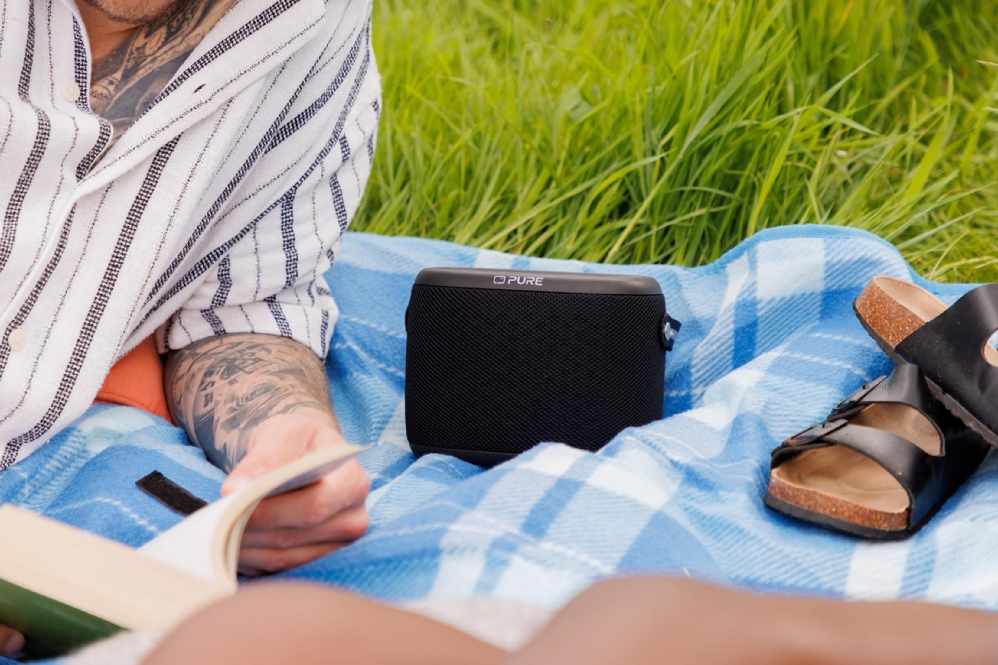 Une personne lit un livre sur une couverture à côté d'un haut-parleur et de sandales.