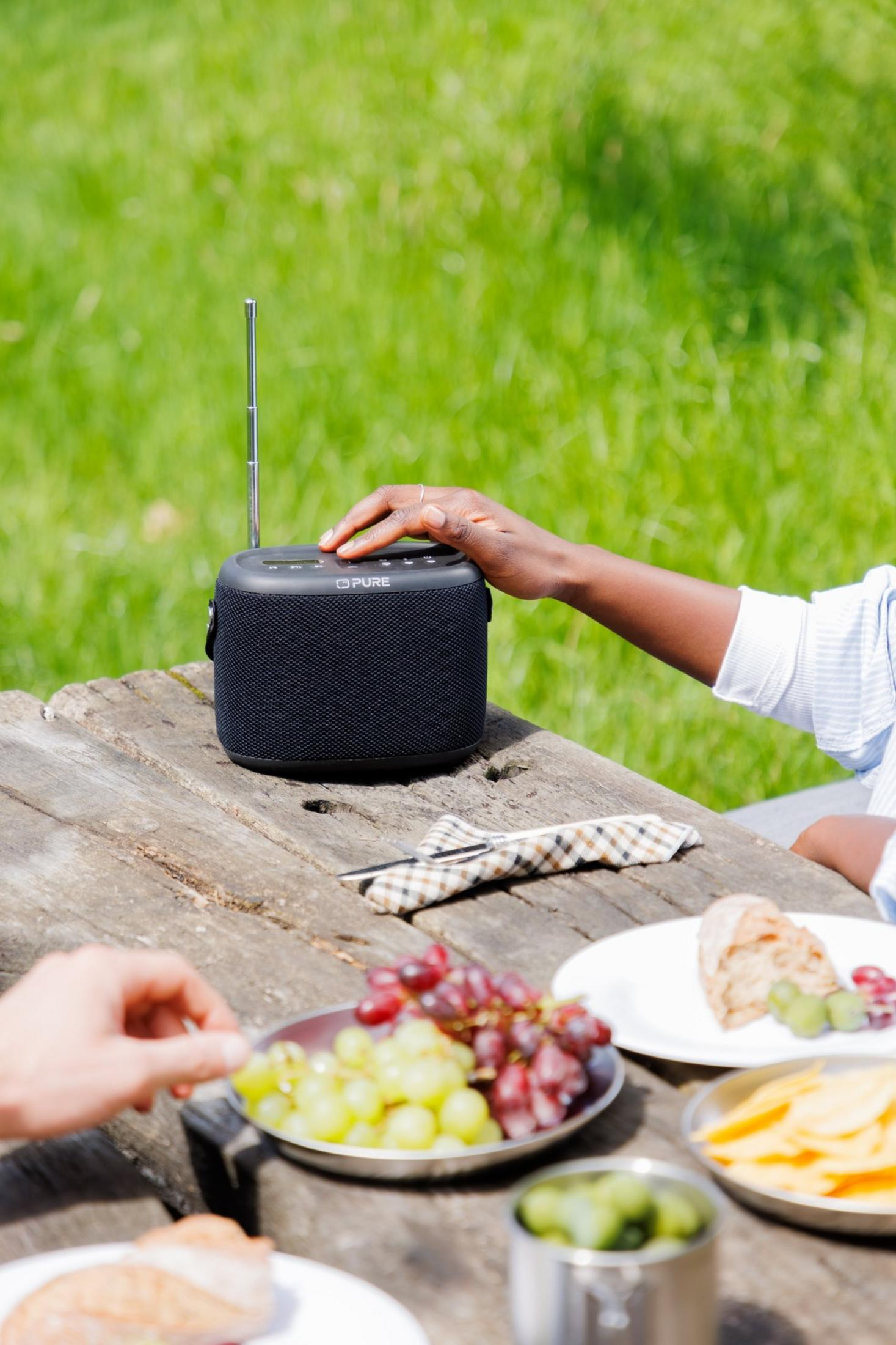 Une enceinte noire sur une table en bois extérieure, avec des fruits et du pain à proximité.