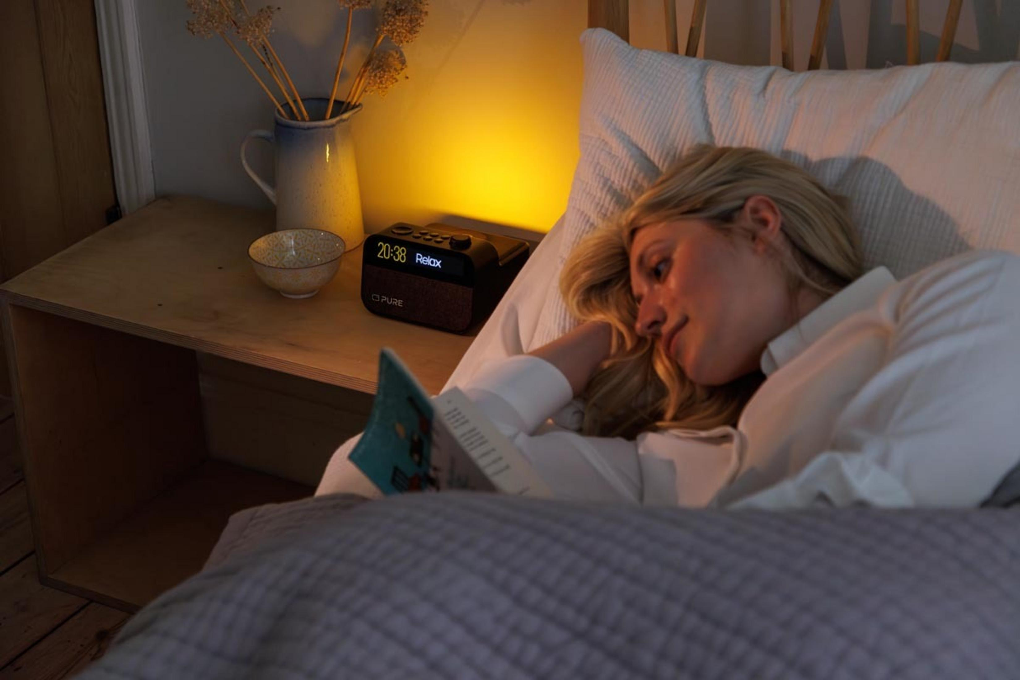 Mujer tumbada en la cama leyendo un libro. Mesita de noche con reloj y jarrón.
