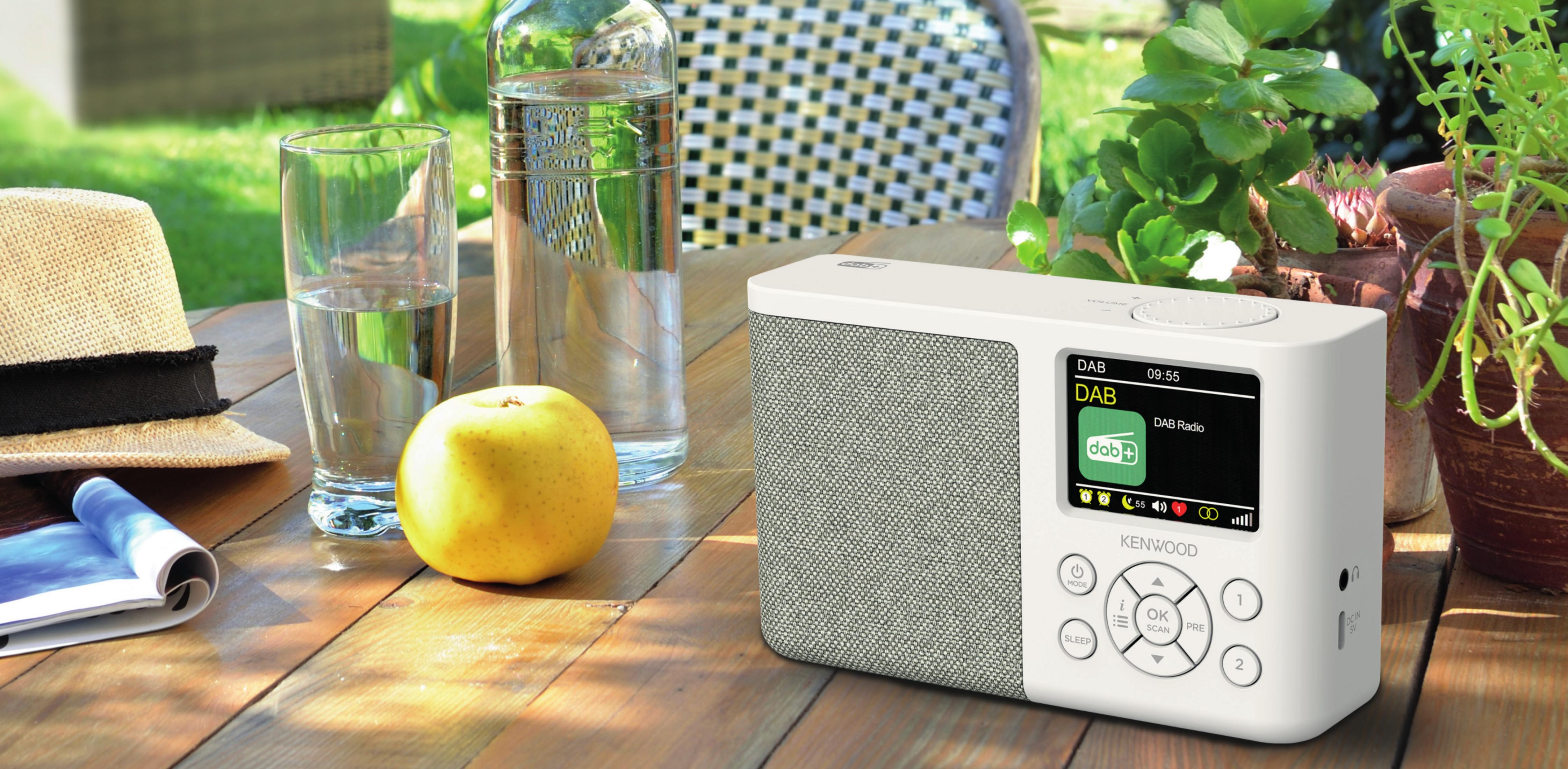 Białe radio DAB stoi na drewnianym stole z kapeluszem, jabłkiem i wodą.