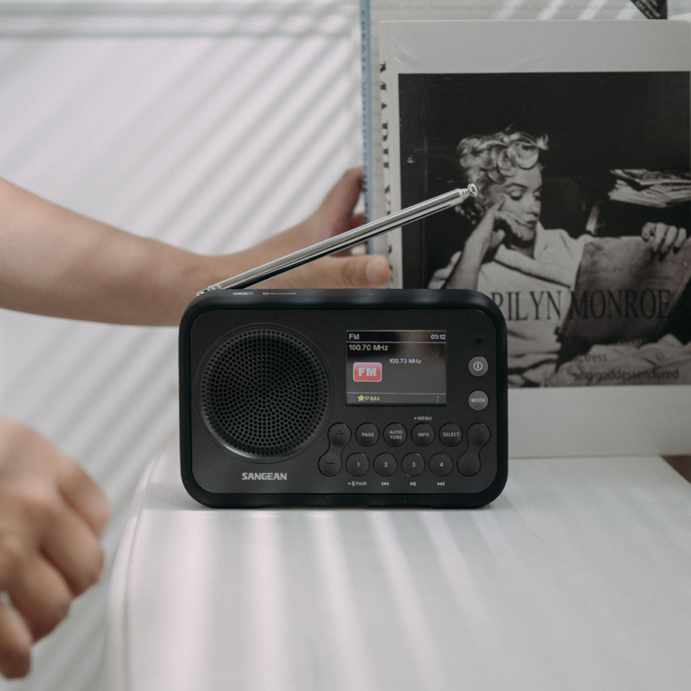 Une radio Sangean noire avec antenne déployée, à côté d'un livre de Marilyn Monroe.