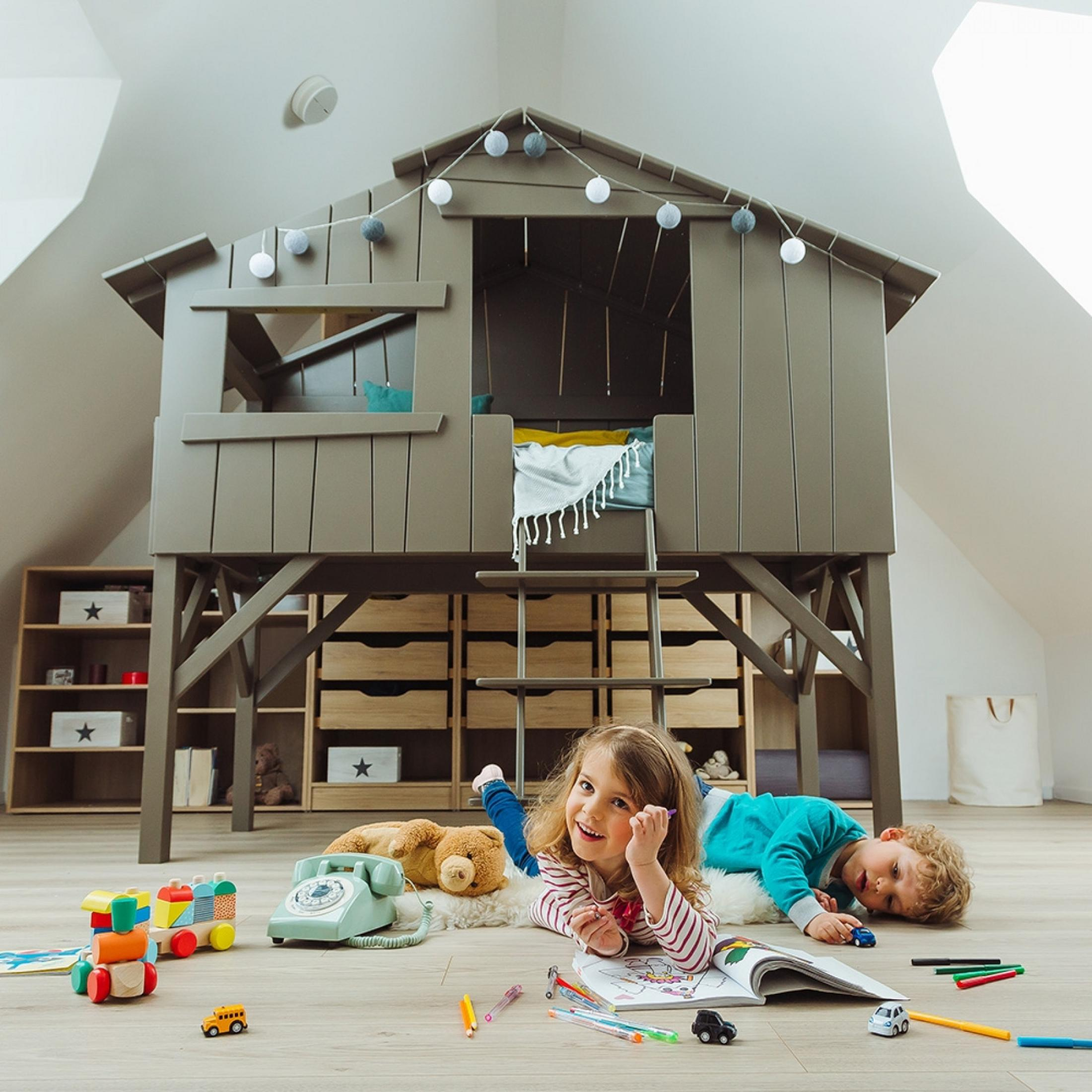 Two children play in front of a playhouse. A girl is drawing, a boy lies nearby.