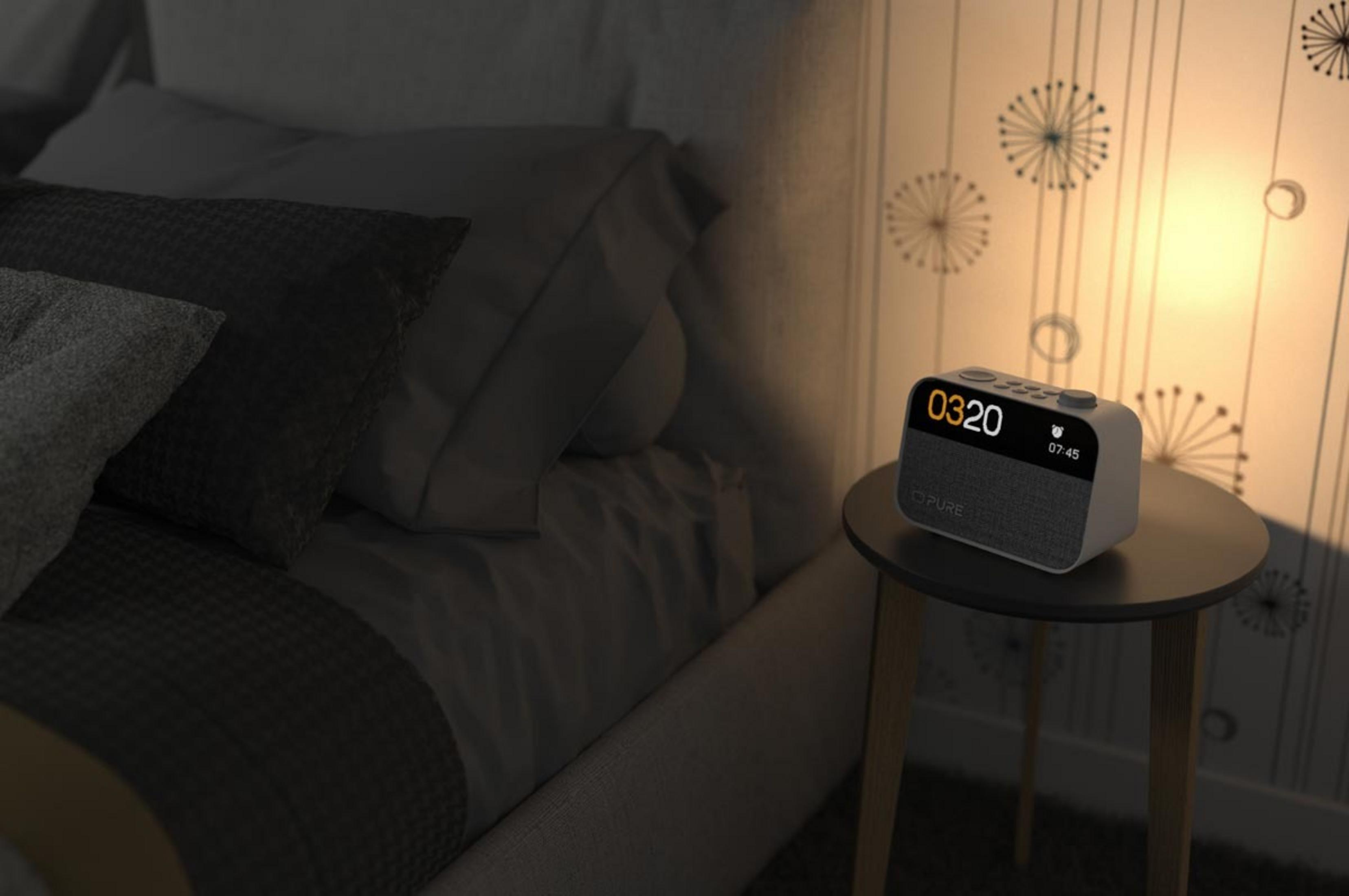 Una mesita de noche con un despertador junto a una cama. La pared tiene un patrón.