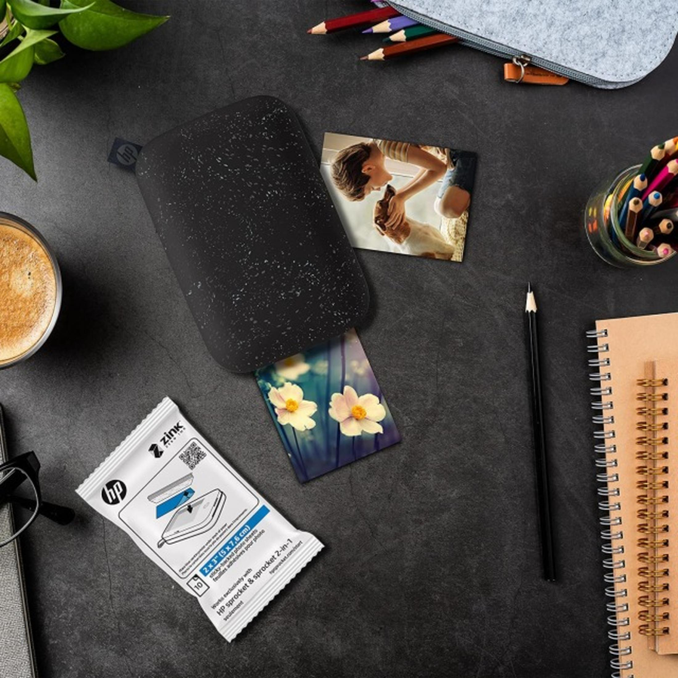 Czarna drukarka HP Sprocket ze zdjęciami, kawą i materiałami plastycznymi.