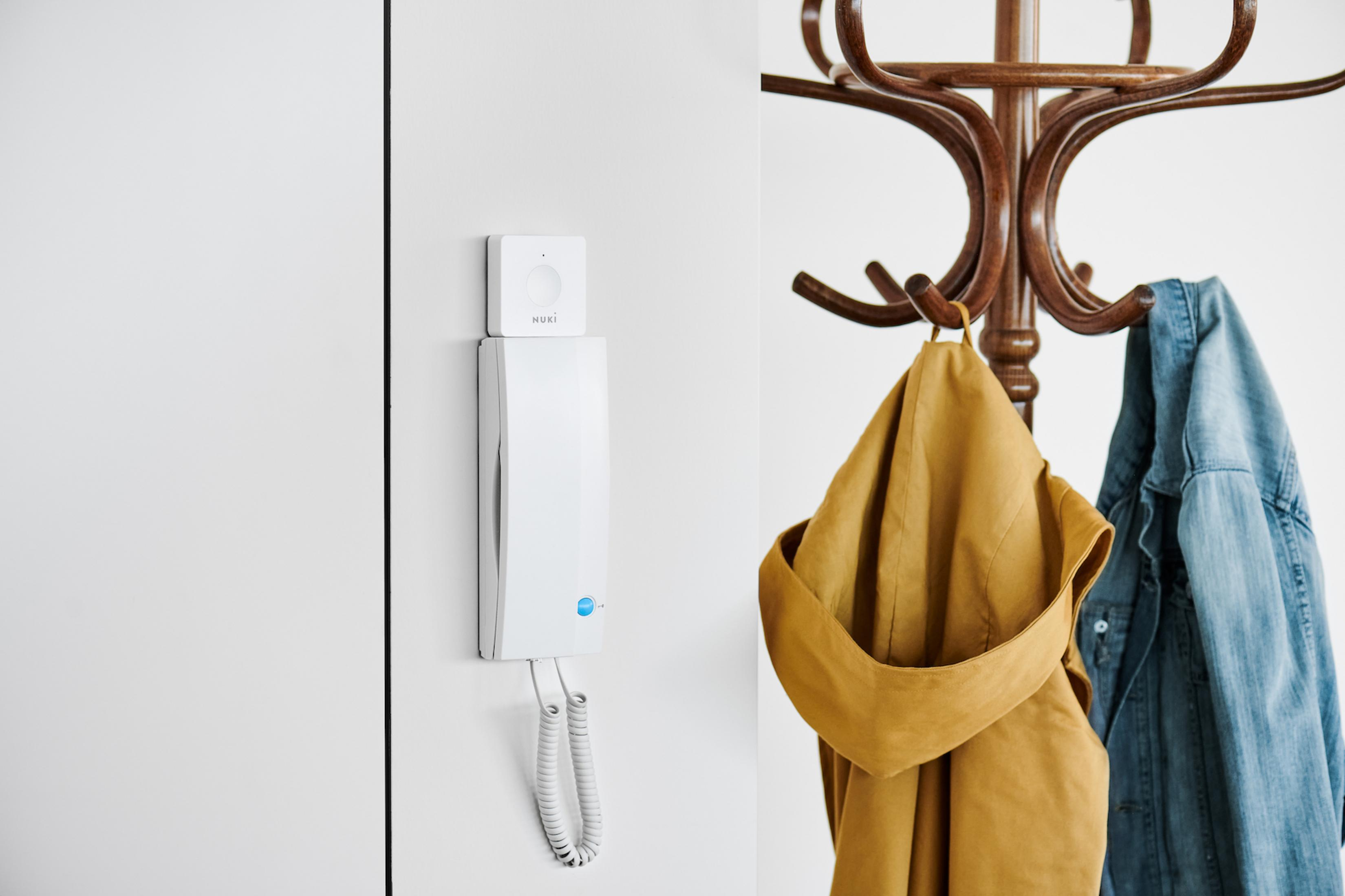 Ein weißes Nuki-Türschloss und eine Gegensprechanlage hängen an der Wand neben einem Holzgarderobenständer mit Kleidung.
