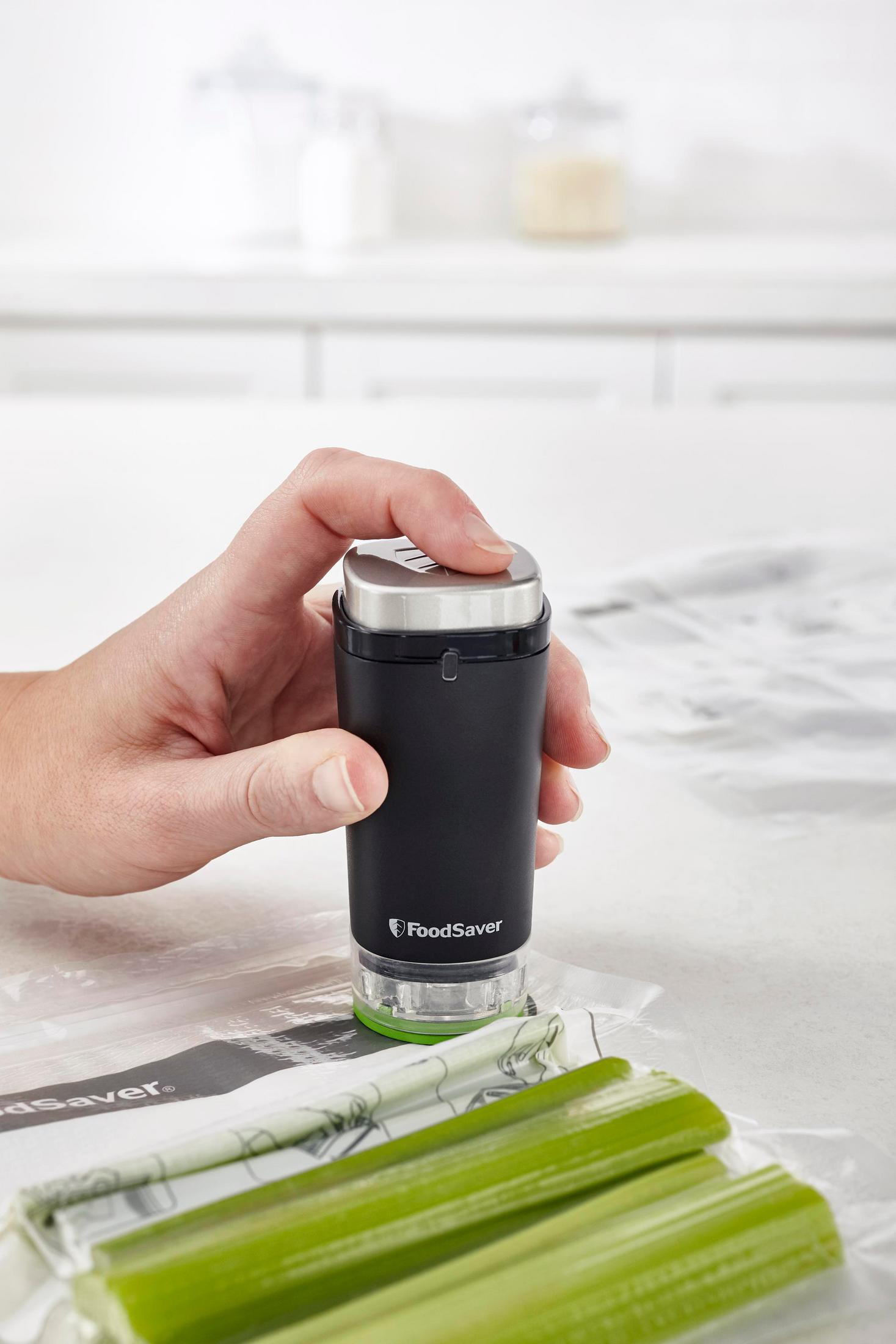 Mano presiona sellador al vacío FoodSaver negro sobre apio en bolsa.