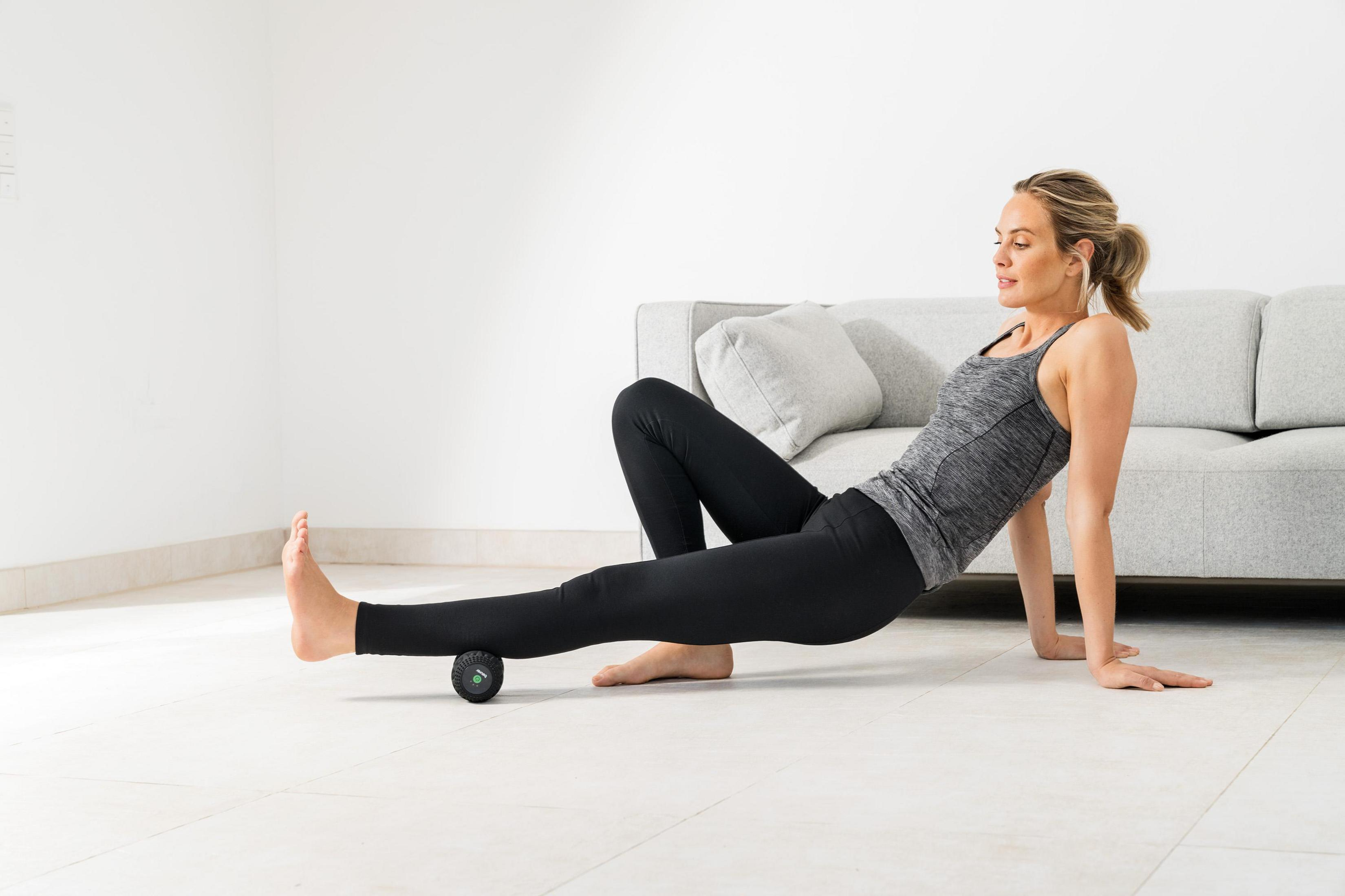 Frau macht Beinmassage mit Faszienrolle auf dem Boden vor Sofa.